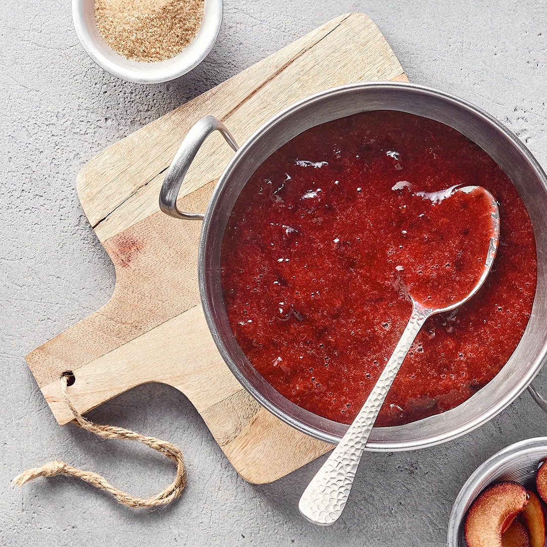Ein Topf mit dicker roter Fruchtsoße, leicht stückig, ein Löffel darin, daneben frische Pflaumen.