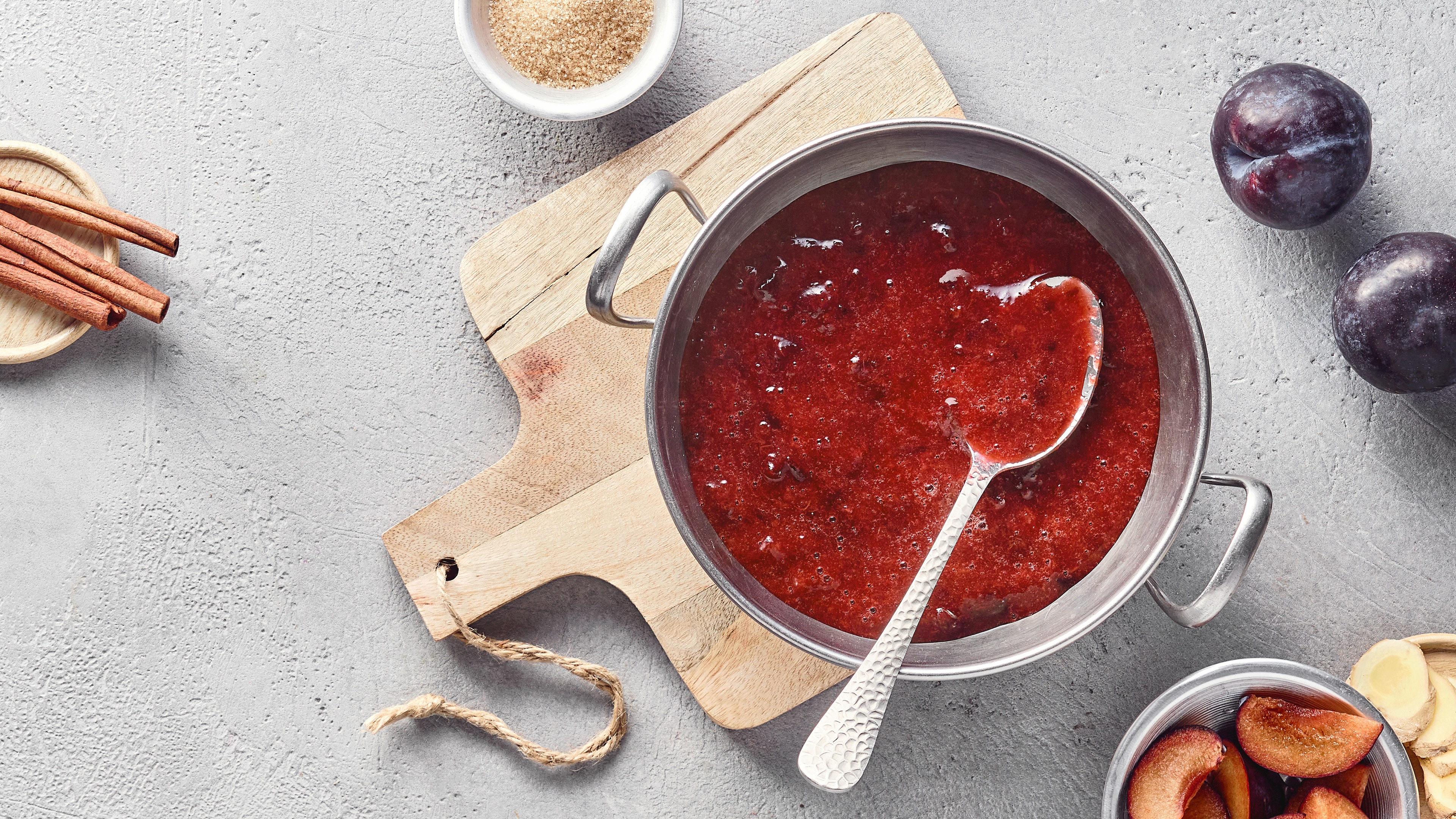 Ein Topf mit dicker roter Fruchtsoße, leicht stückig, ein Löffel darin, daneben frische Pflaumen.