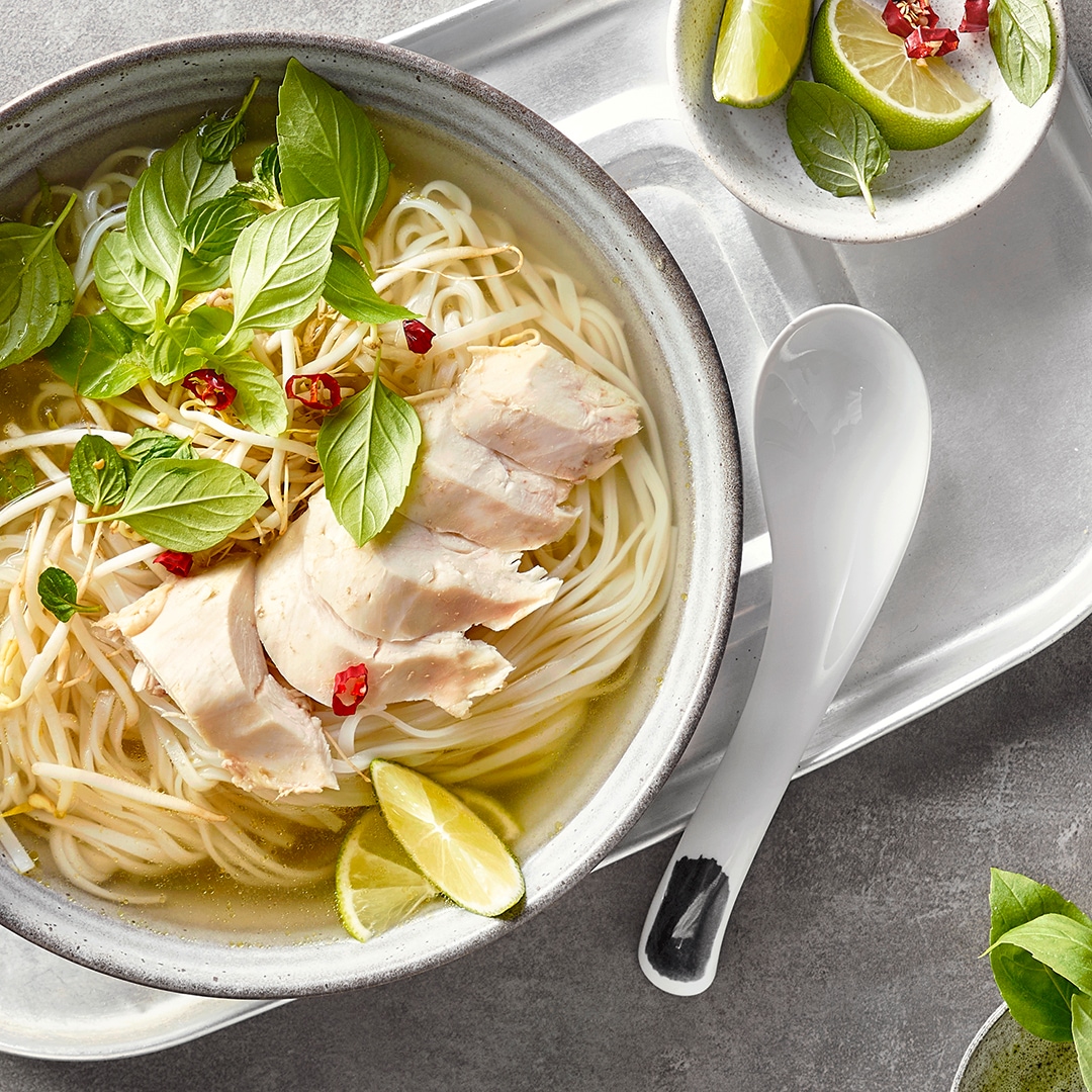 Eine Schale mit klarer Brühe, langen Nudeln, zarten Hühnerstreifen, garniert mit frischen Basilikumblättern, roten Chilistücken & Limettenspalten