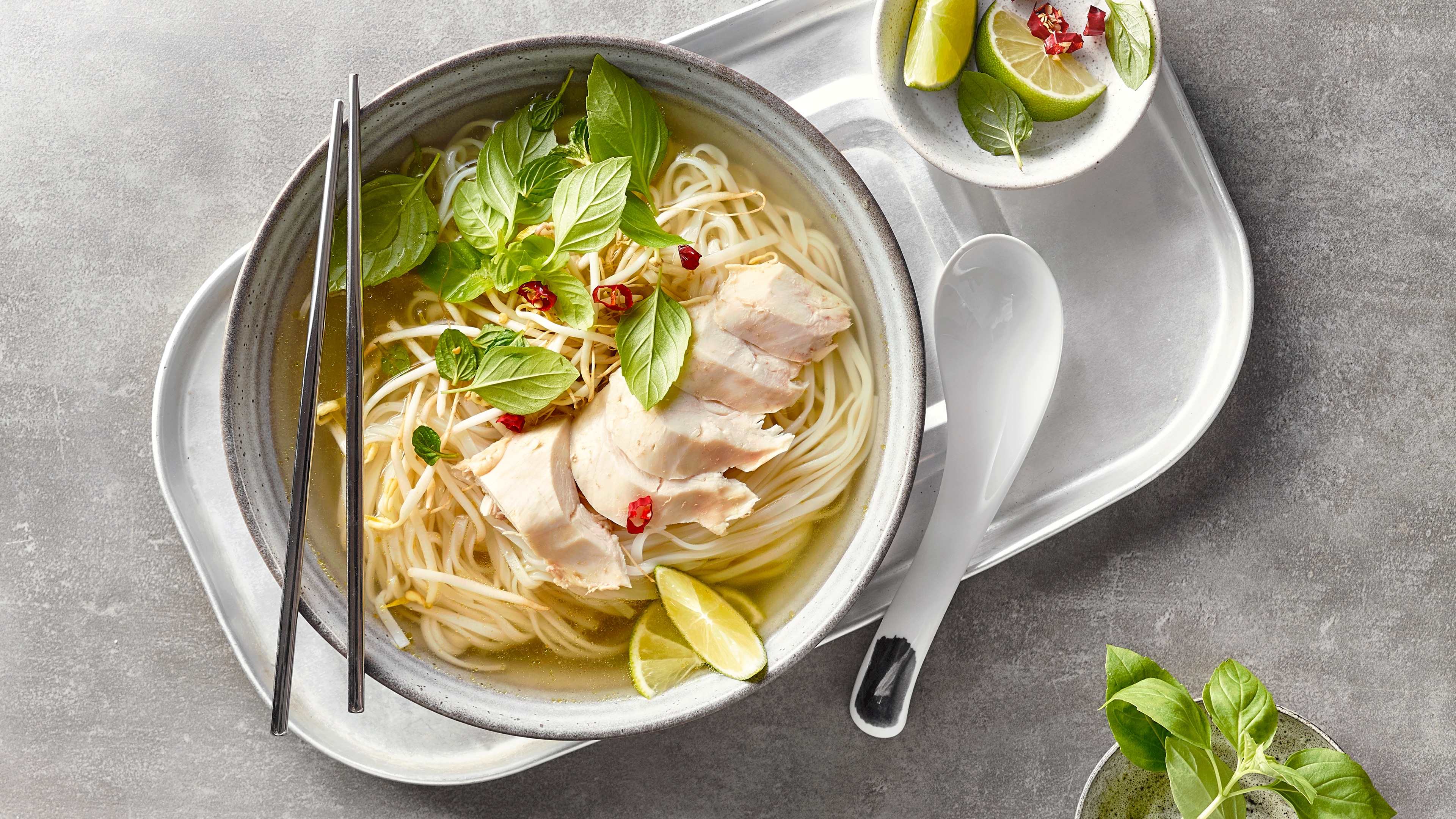 Eine Schale mit klarer Brühe, langen Nudeln, zarten Hühnerstreifen, garniert mit frischen Basilikumblättern, roten Chilistücken und Limettenspalten
