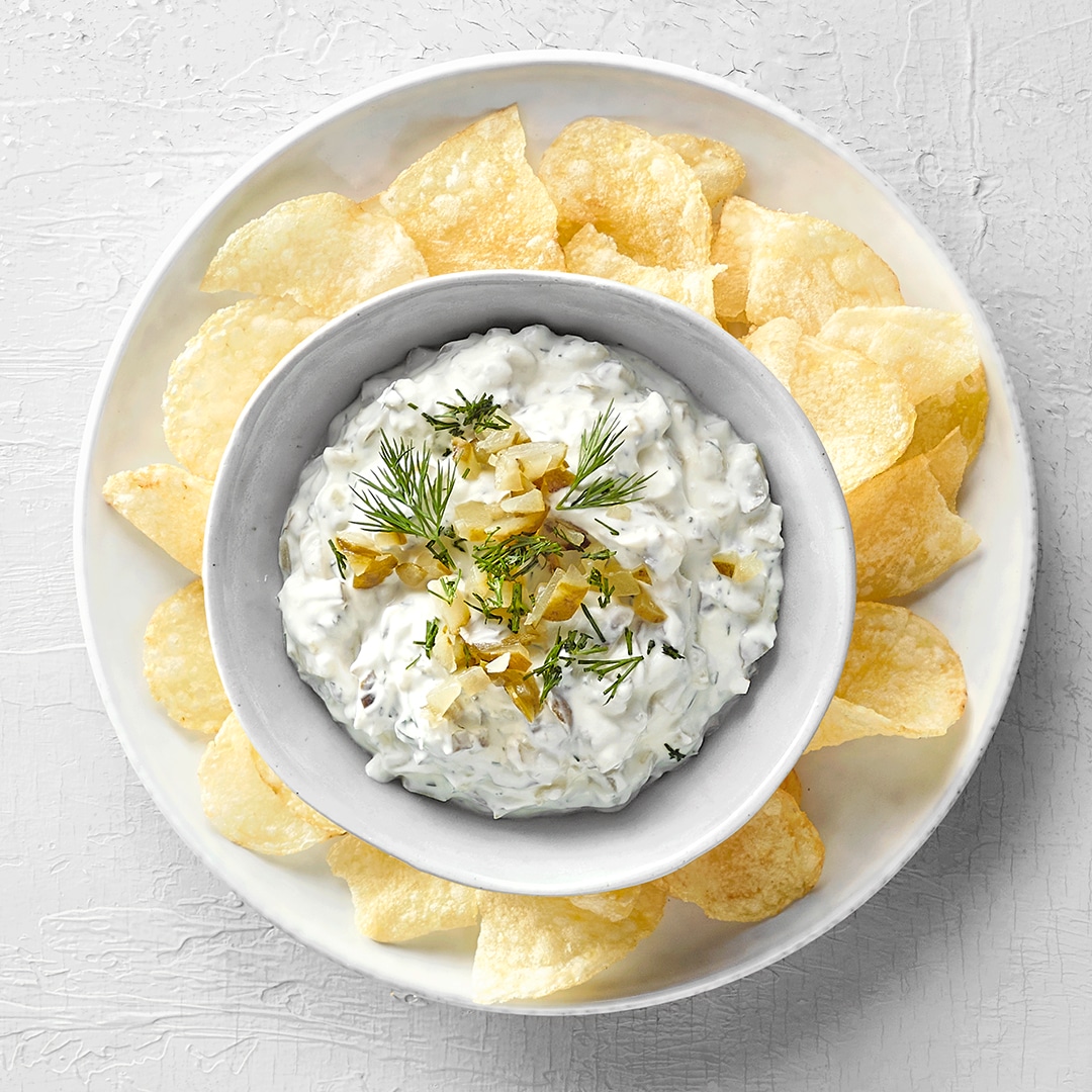 Schale mit Kartoffelchips. In der Mitte der Schale steht eine kleinere Schale mit Dip, zerstückelten Gewürzgurken und Rosmarin.