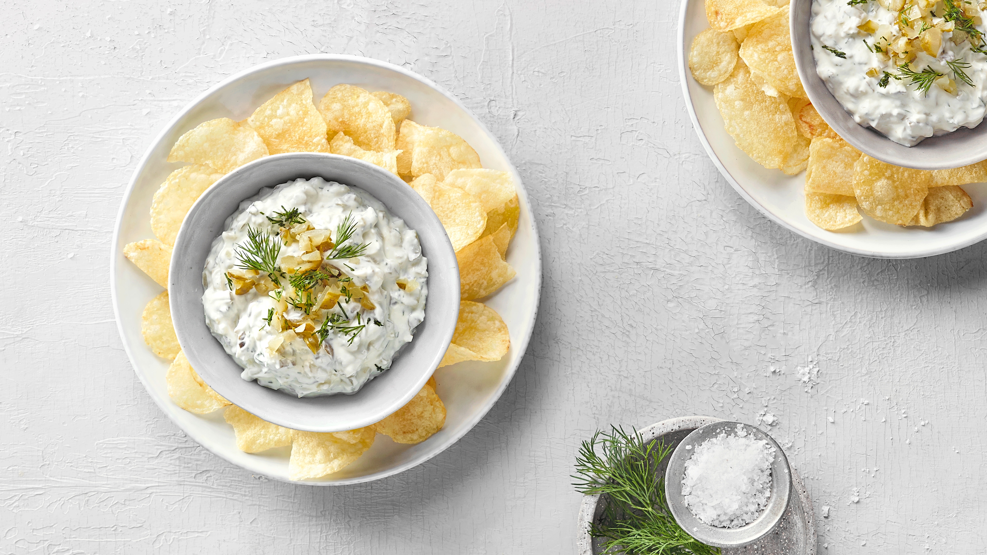Zwei Schalen mit Kartoffelchips. In der Mitte der Schale steht jeweils eine kleinere Schale mit Dip, zerstückelten Gewürzgurken und Rosmarin.