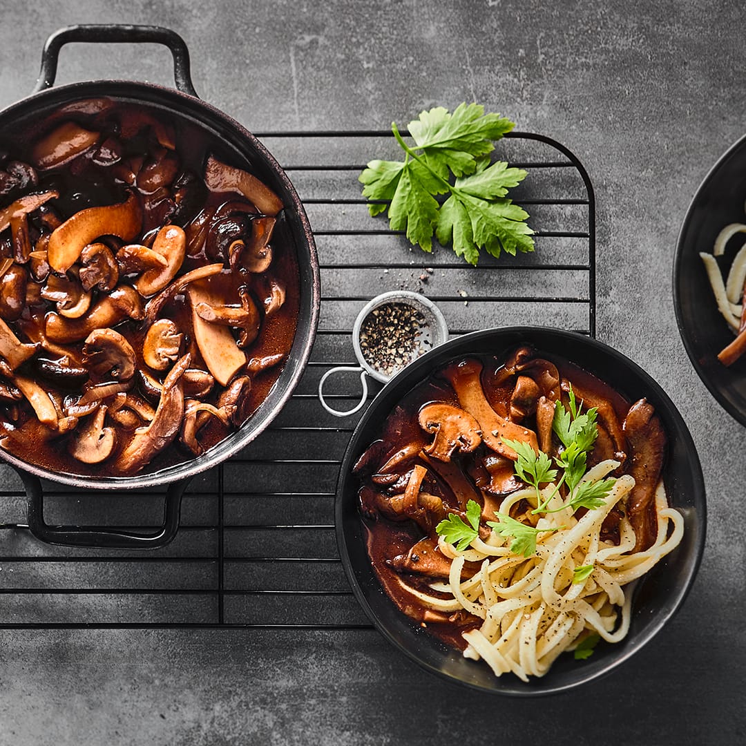 Eintopfgericht aus Champignons und Kräuterseitlingen in dunkler Soße, getoppt mit Petersilie, in einem gusseisernen Topf.
