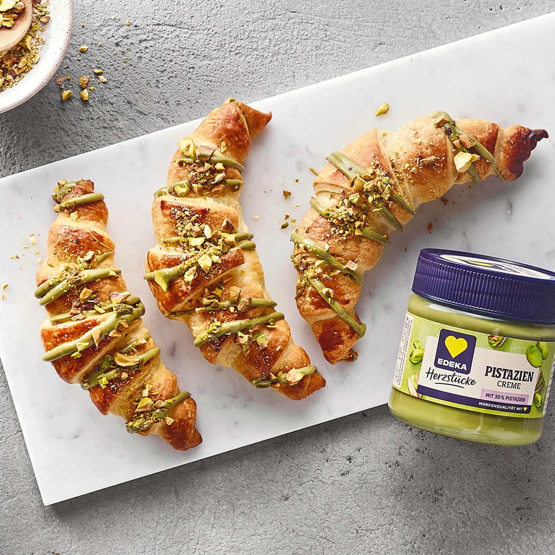 Buttercroissants mit Pistaziencreme‑Füllung, bestreut mit gehackten Pistazien. Daneben EDEKA Herzstücke Pistaziencreme.
