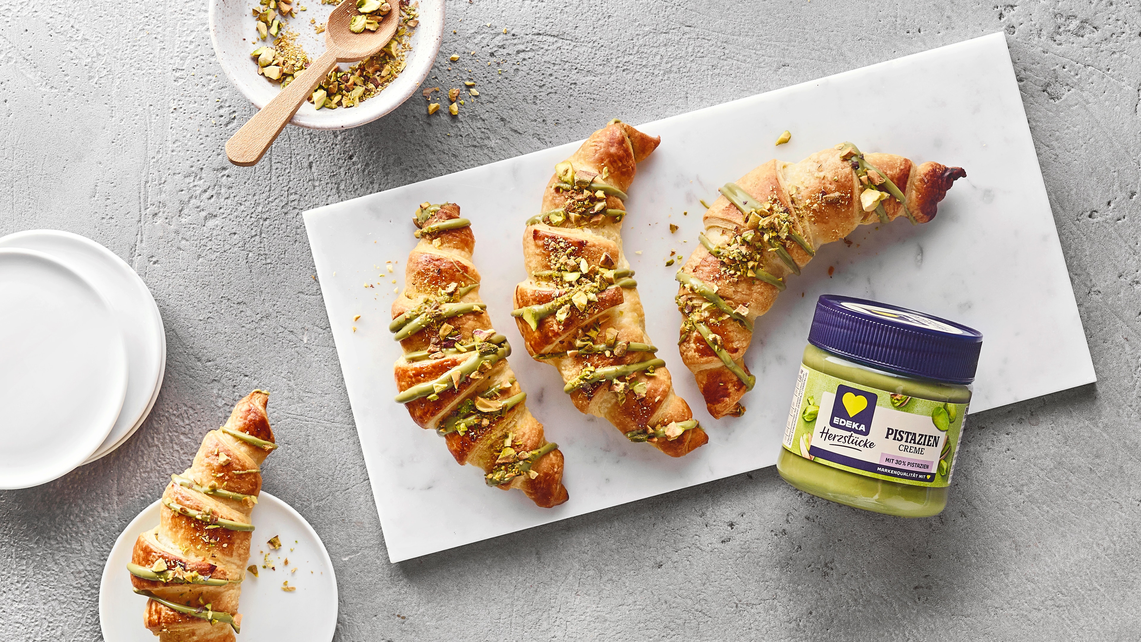 Buttercroissants mit Pistaziencreme‑Füllung, bestreut mit gehackten Pistazien. Daneben EDEKA Herzstücke Pistaziencreme.