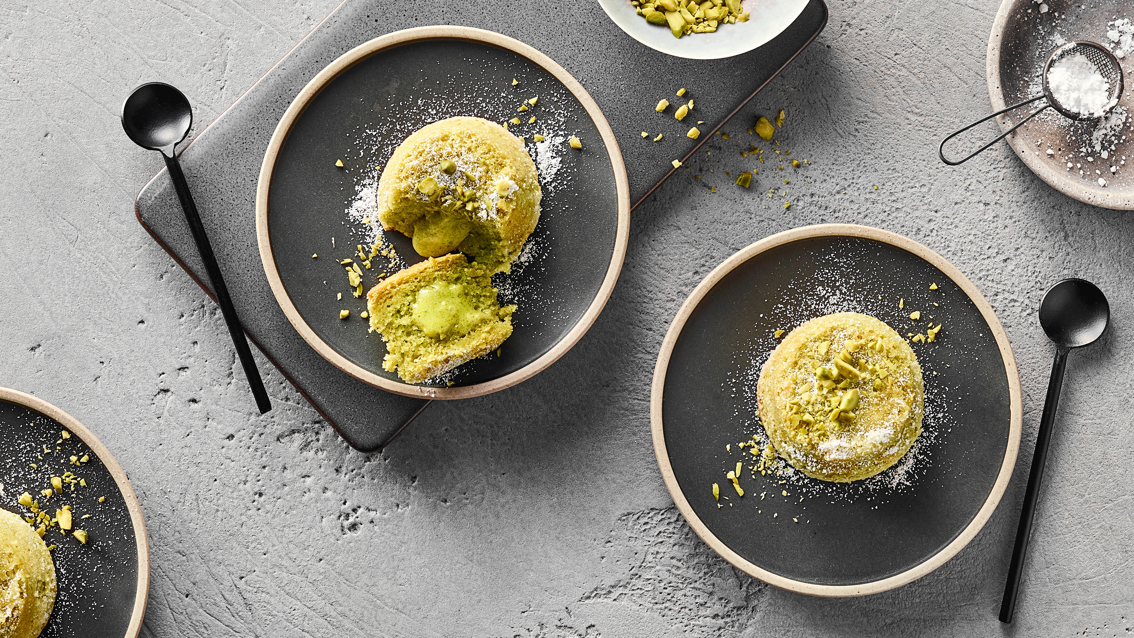 Rezept für Pistazien-Soufflé Ein Teller mit einem grünen Törtchen mit einem flüssigen Kern garniert mit Pistazienstücken und weißen Raspeln
