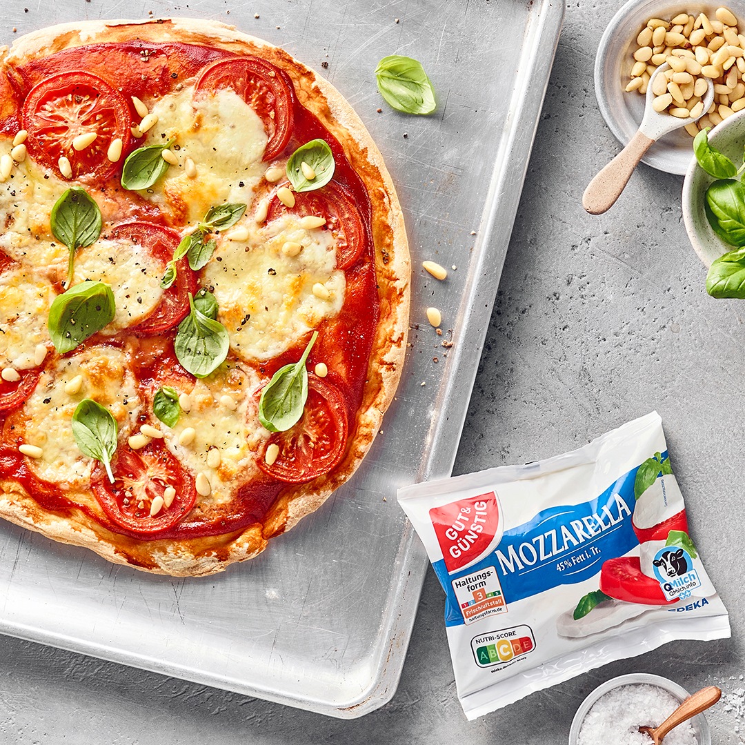 Pizza mit Tomaten, Mozzarella und frischem Basilikum auf einem Backblech, daneben Pinienkerne und GUT&GÜNSTIG Mozzarella.
