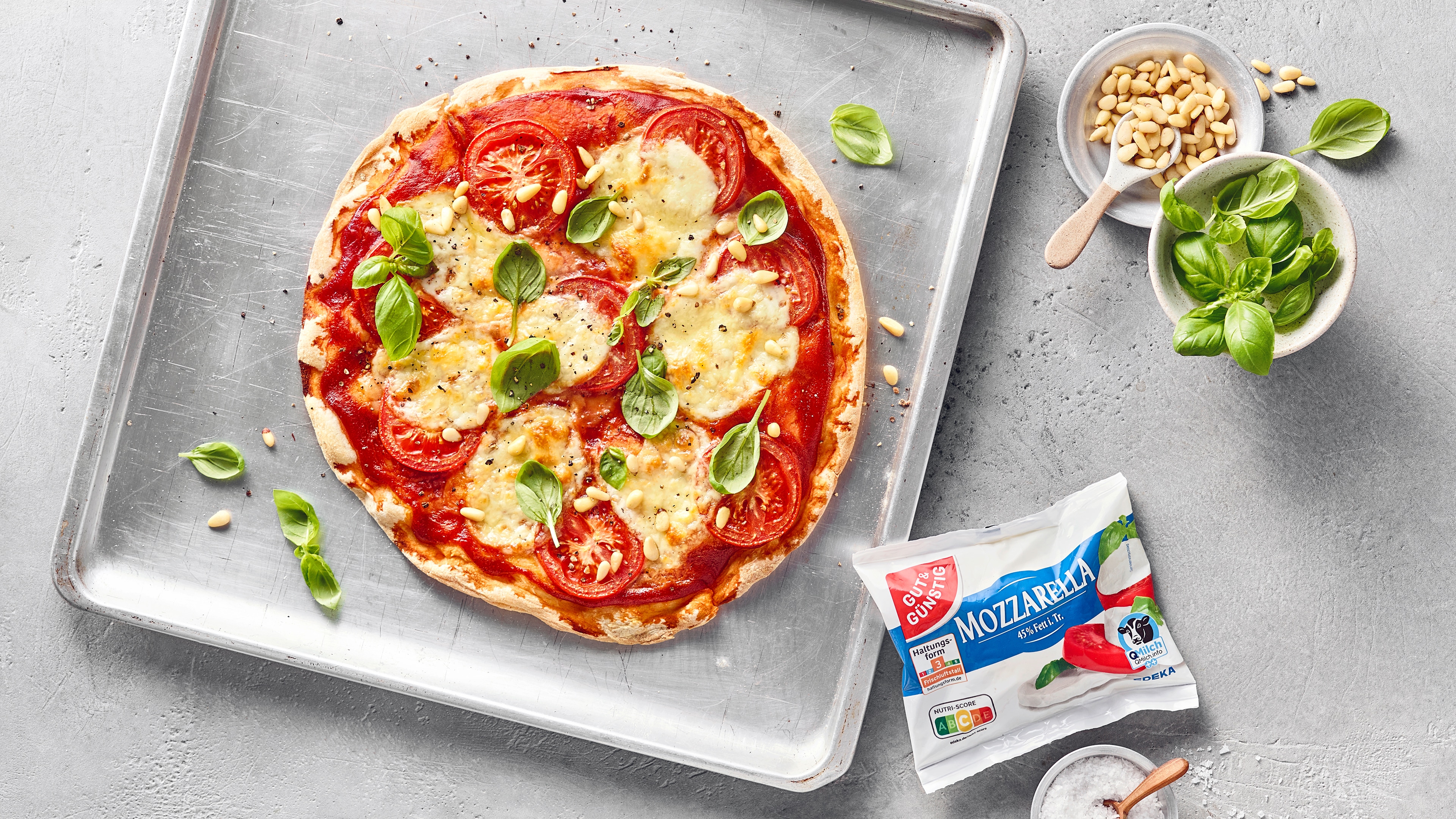 Pizza mit Tomaten, Mozzarella und frischem Basilikum auf einem Backblech, daneben Pinienkerne und GUT&GÜNSTIG Mozzarella.