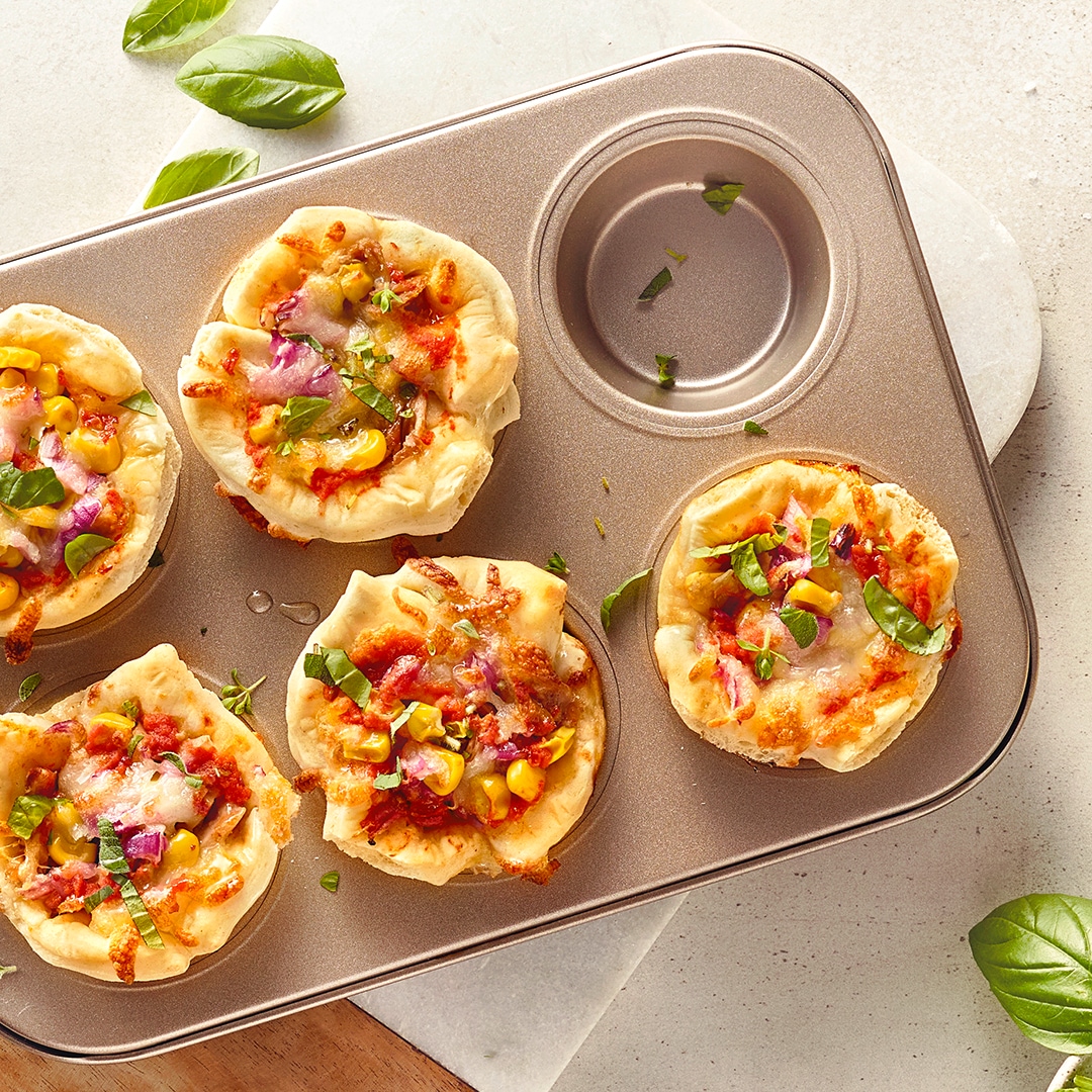 Ein Brett mit einem Muffin aus Pizzateig, darin Tomaten und Mais, graniert mit Basilikum