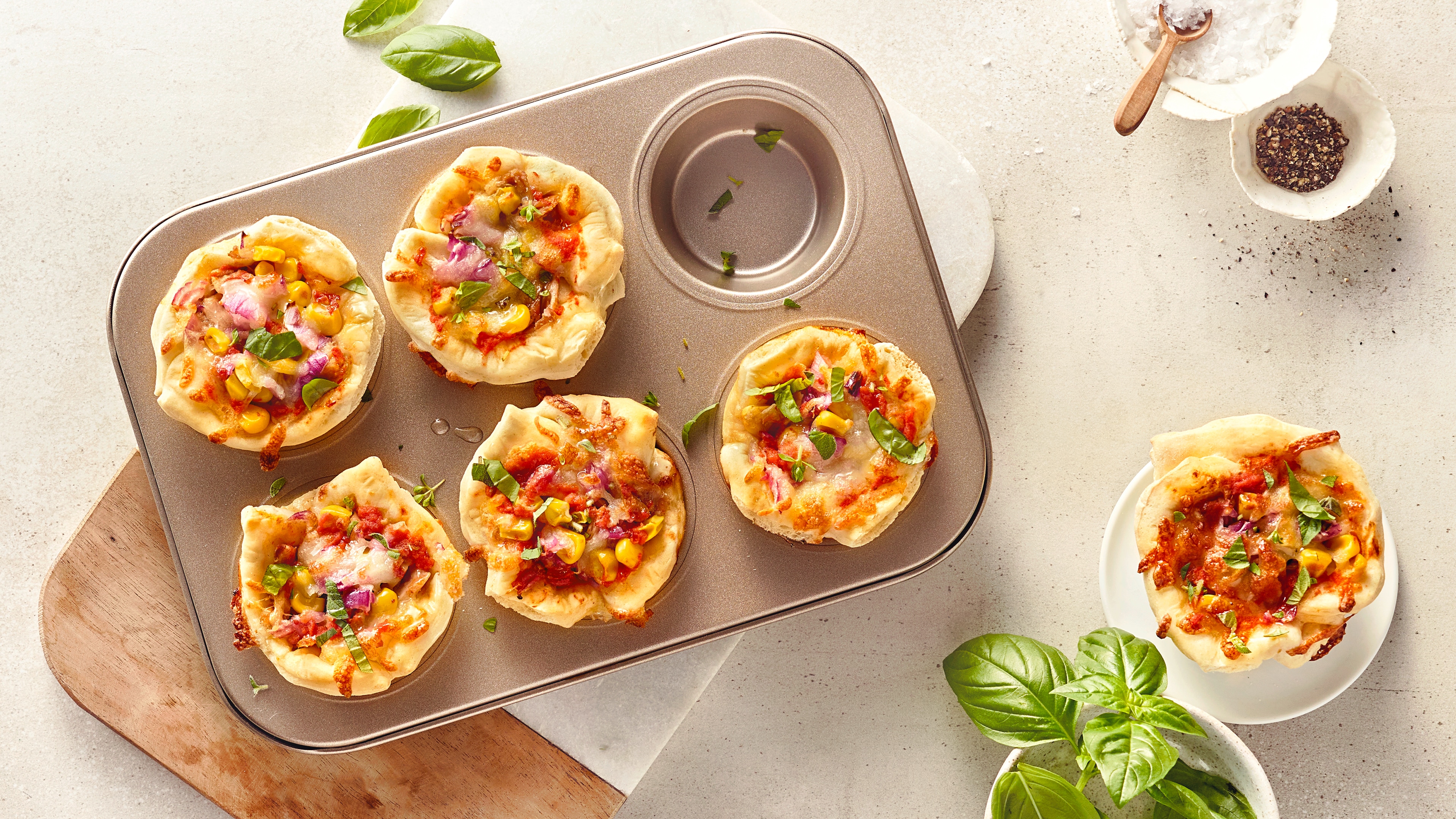 Ein Brett mit einem Muffin aus Pizzateig, darin Tomaten und Mais, graniert mit Basilikum