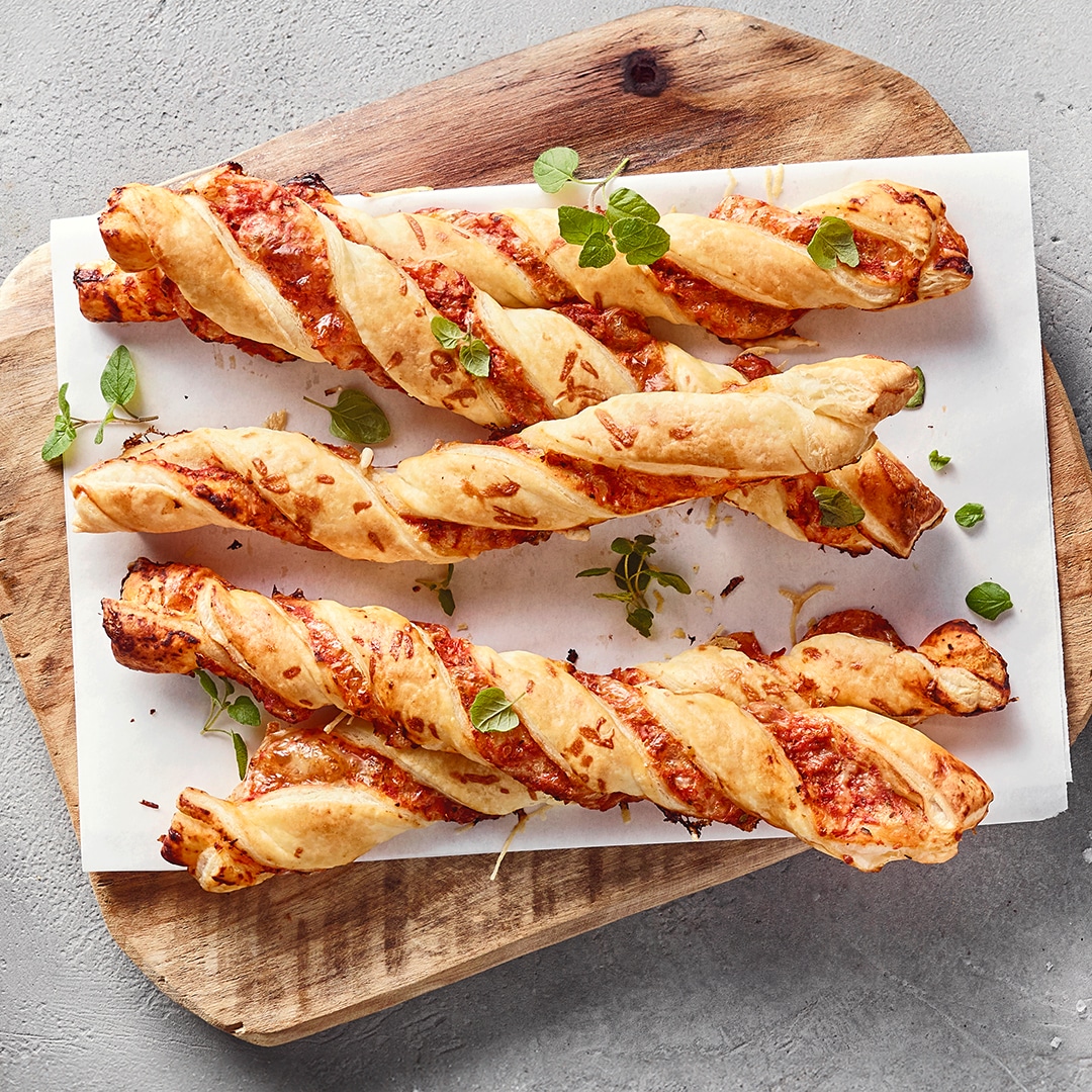 Ein Brett mit gedrehten hellbraunen Teigstangen, gefüllt mit Tomatensauce und garniert mit frischen Kräutern.