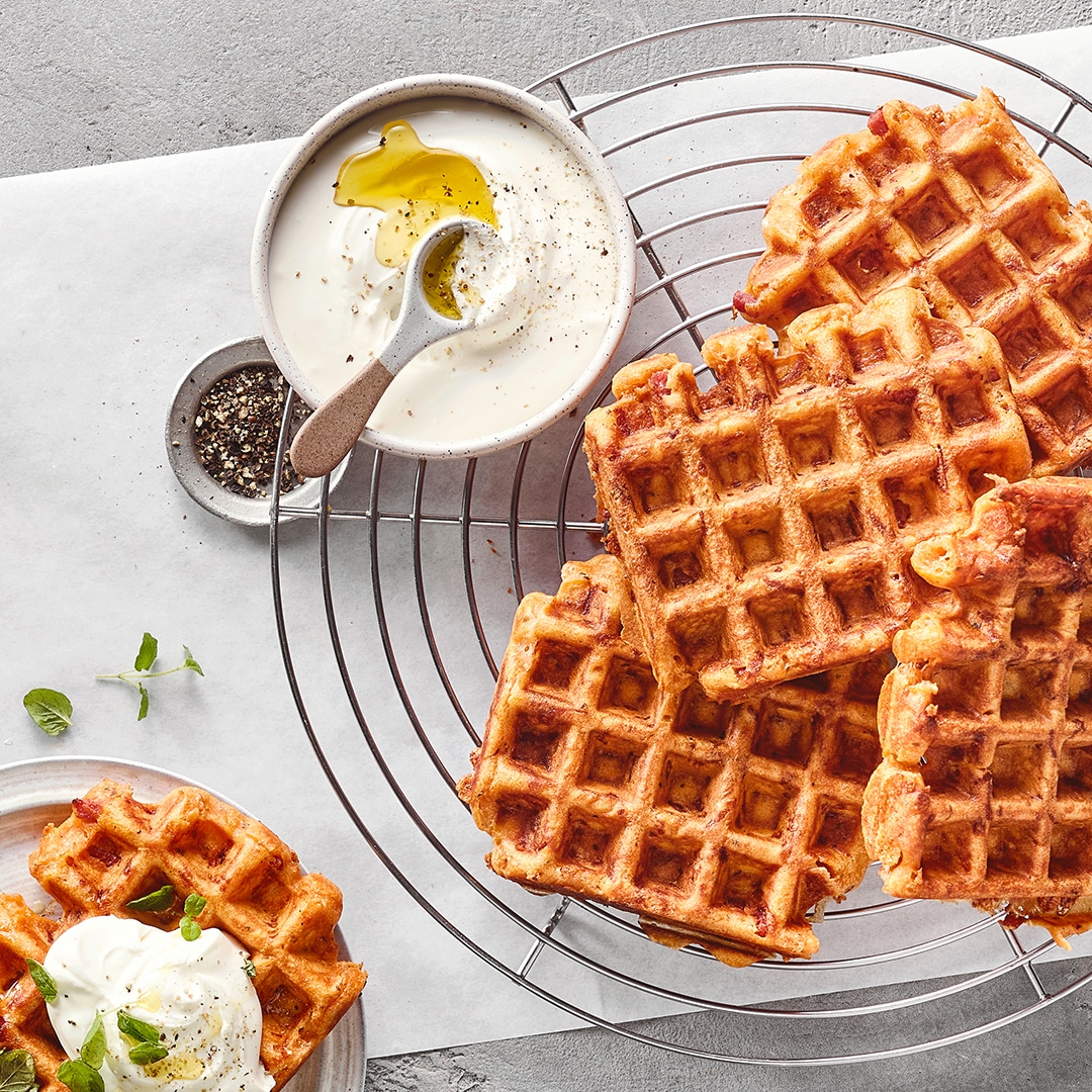 Ein Gitter mit goldbraunen Waffeln, daneben eine Schüssel mit weißer Creme, Öl und Pfeffer sowie Kräutern auf den Waffeln.