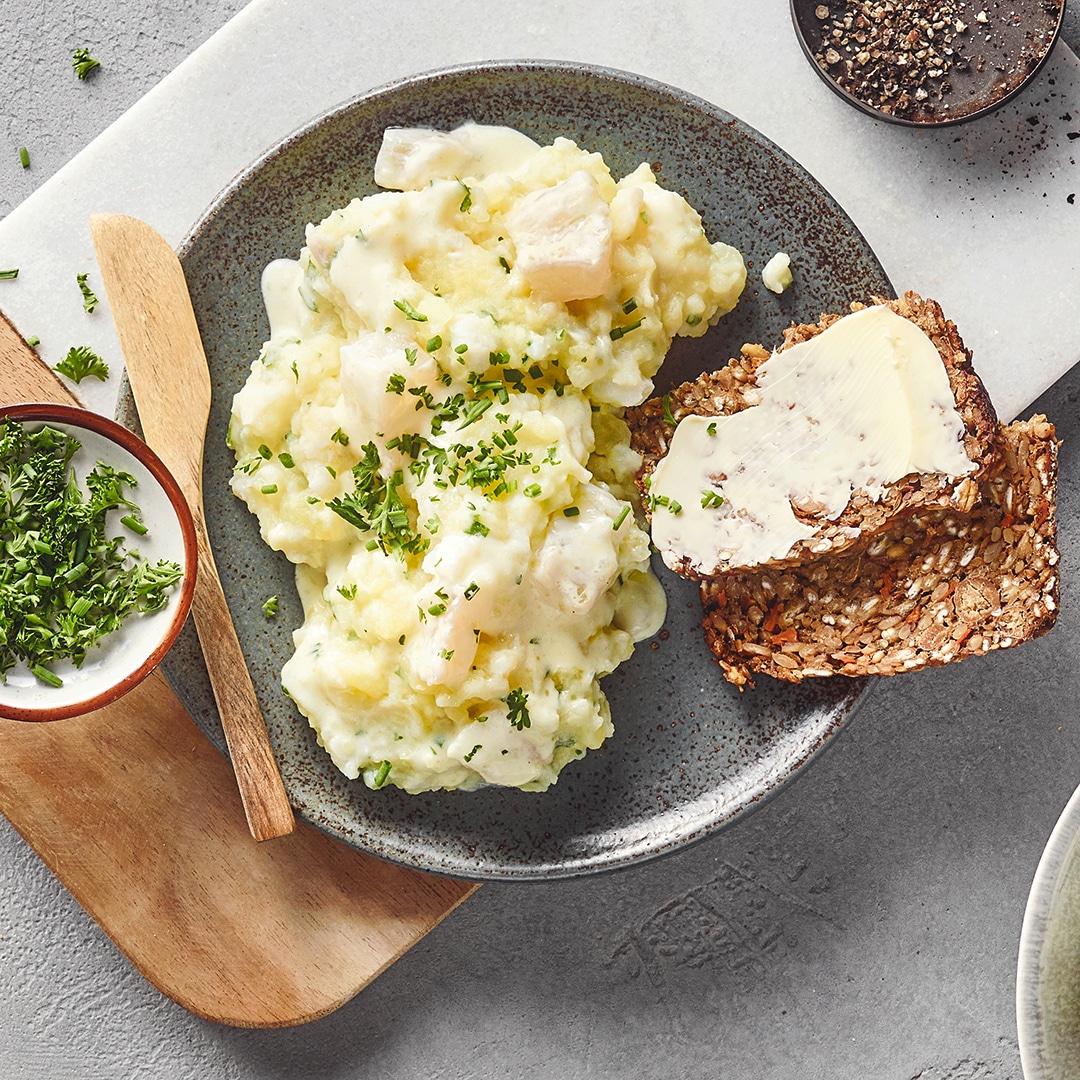 Ein Teller mit einem weißen, cremigen Brei mit grünen Kräutern, daneben zwei Scheiben braunes Körnerbrot mit Butter.