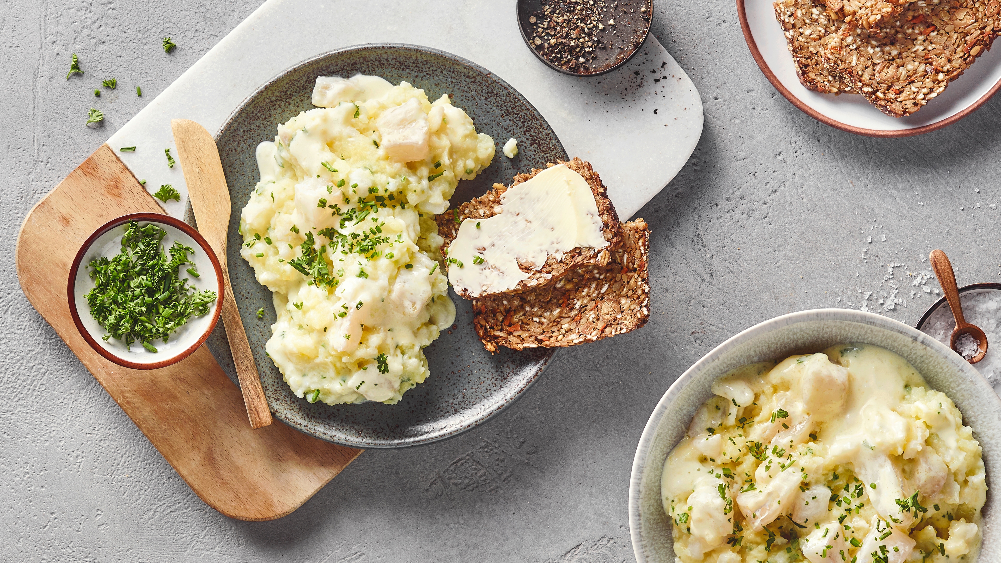 Ein Teller mit einem weißen, cremigen Brei mit grünen Kräutern, daneben zwei Scheiben braunes Körnerbrot mit Butter.