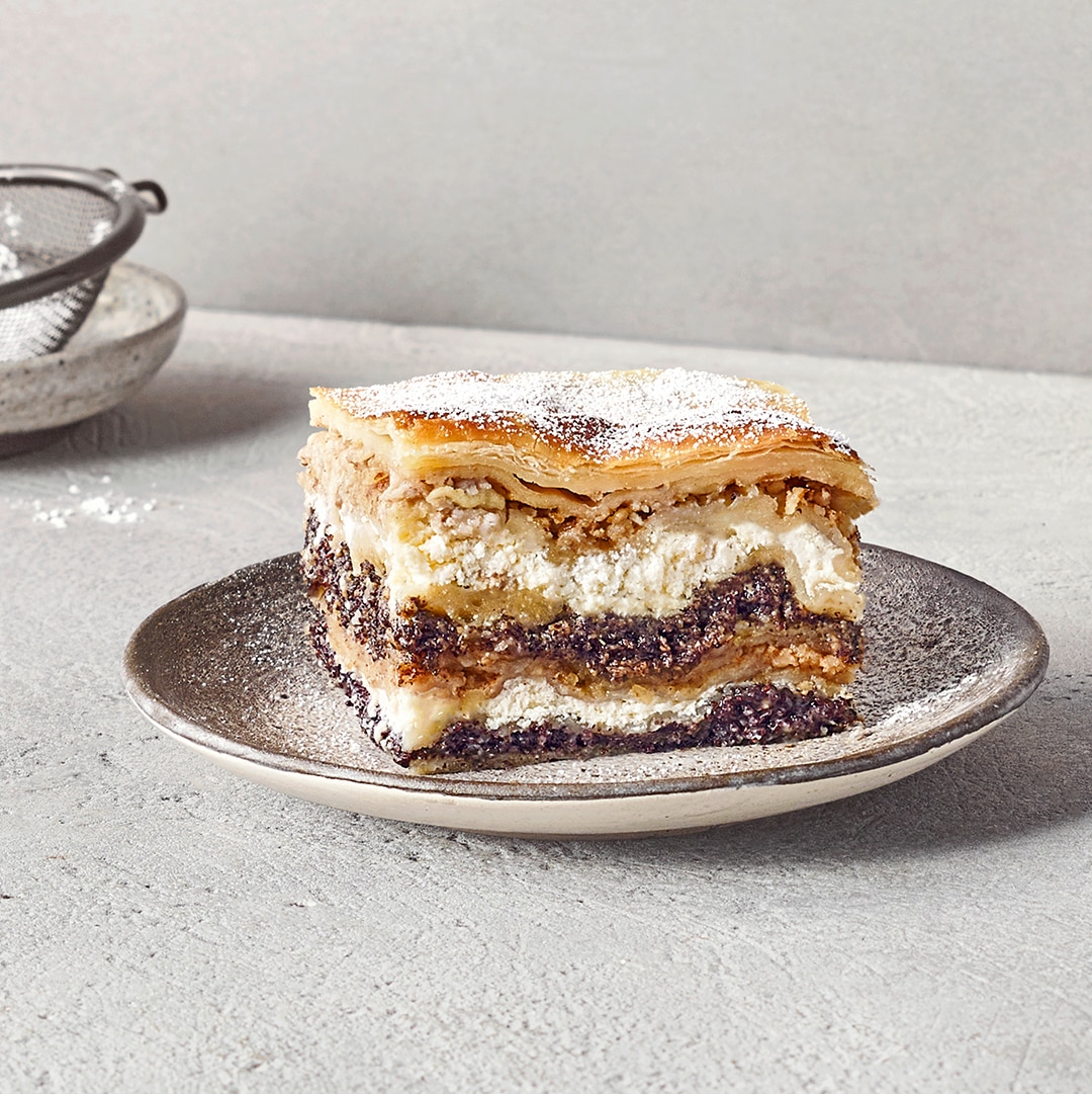 Ein Teller mit einem Stück Schichtkuchen aus hellen Teiglagen, weißer Füllung und dunkler Mohnschicht, bestäubt mit Puderzucker.