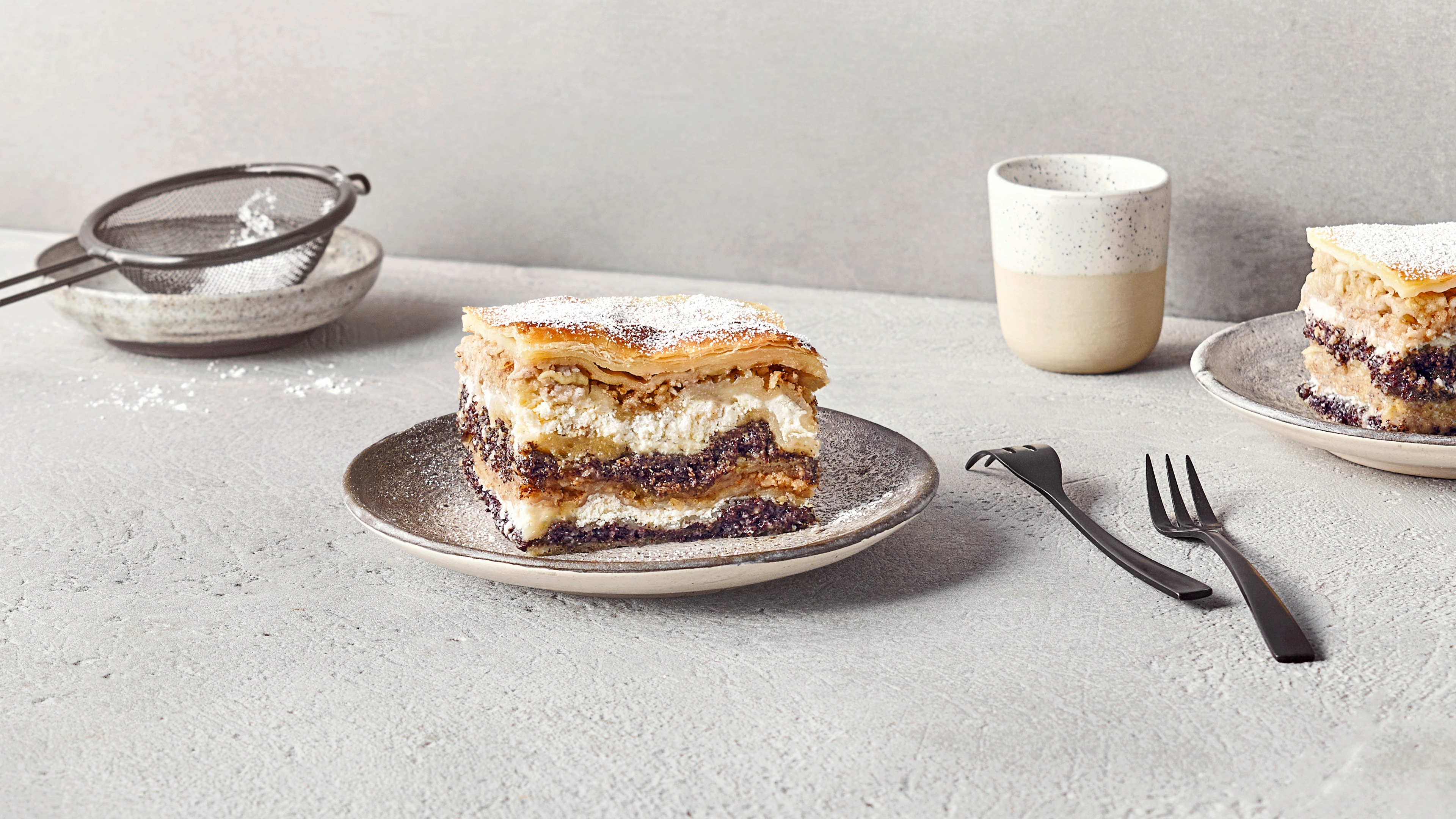 Ein Teller mit einem Stück Schichtkuchen aus hellen Teiglagen, weißer Füllung und dunkler Mohnschicht, bestäubt mit Puderzucker.