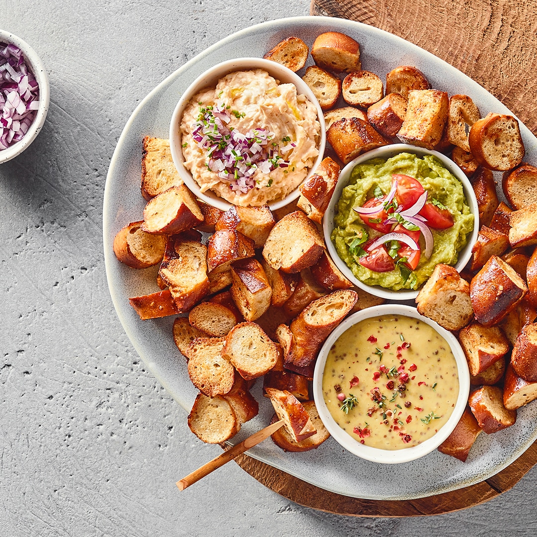 Ein Teller mit braunen Brotstücken und drei Schalen: eine mit heller Creme und Zwiebelstücken, eine mit grünem Dip und Tomaten, eine mit gelbem Dip und Kräutern