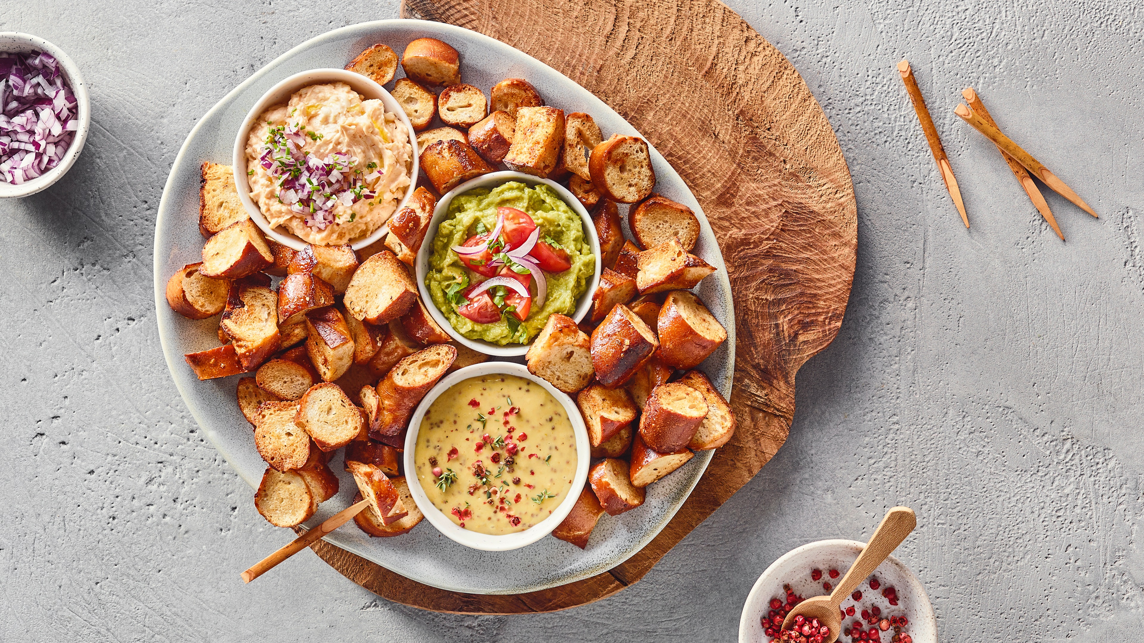 Ein Teller mit braunen Brotstücken und drei Schalen: eine mit heller Creme und Zwiebelstücken, eine mit grünem Dip und Tomaten, eine mit gelbem Dip und Kräutern