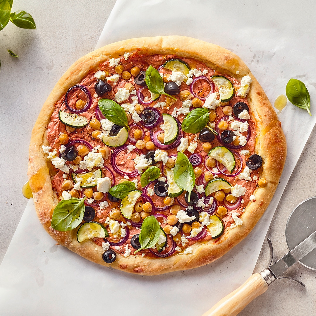 Eine Pizza mit gelbgold gebackenem Rand, hellroter Sauce, Zucchini, Kichererbsen, roter Zwiebel, Oliven, Feta und Basilikum