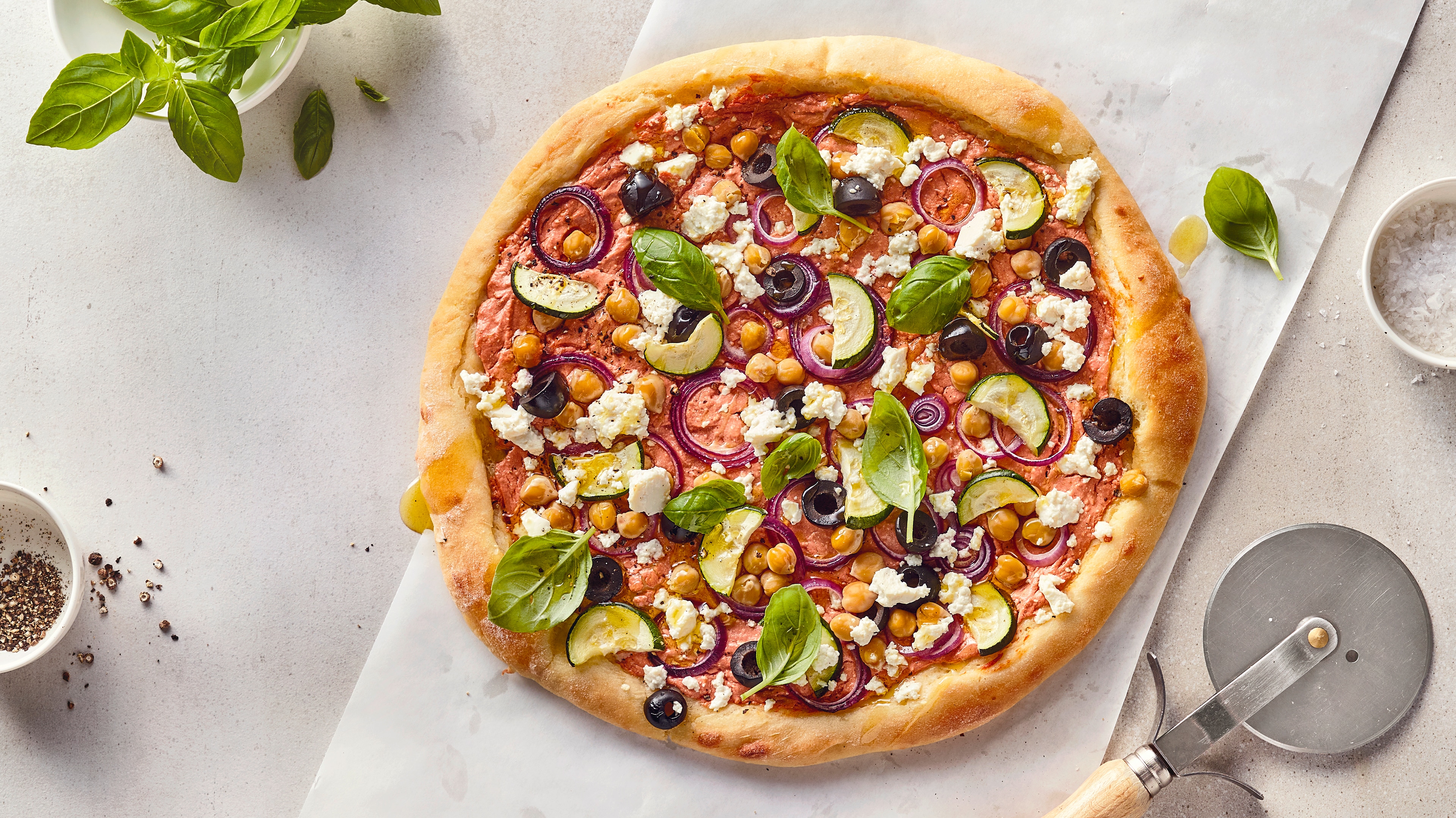 Rezept für Protein-Pizza mit Magerquark Eine Pizza mit gelbgold gebackenem Rand, hellroter Sauce, Zucchini, Kichererbsen, roter Zwiebel, Oliven, Feta und Basilikum