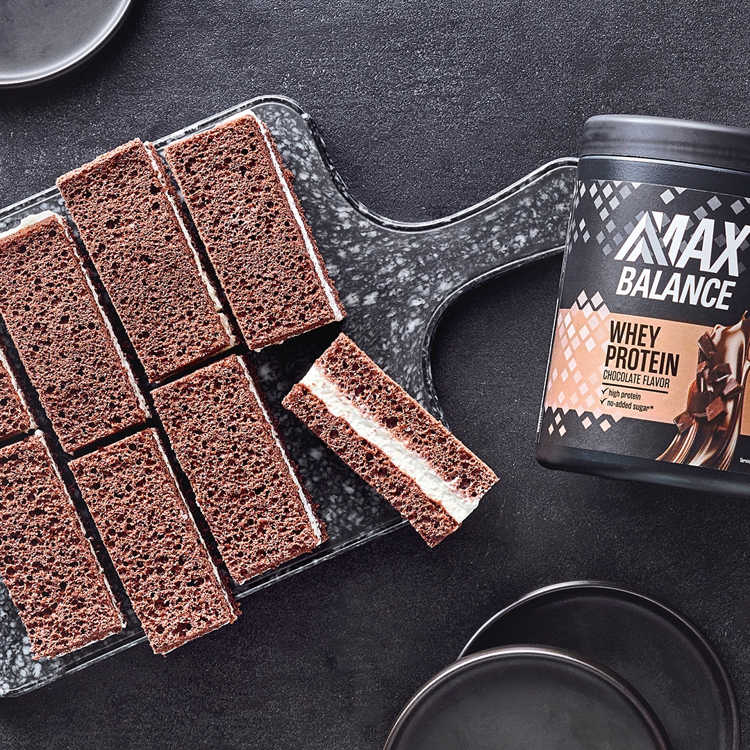 Teigige mit heller Creme gefüllte dunkle Schnitten auf Brettchen, daneben eine Dose MaxBalance Whey Proteinpulver Schokolade