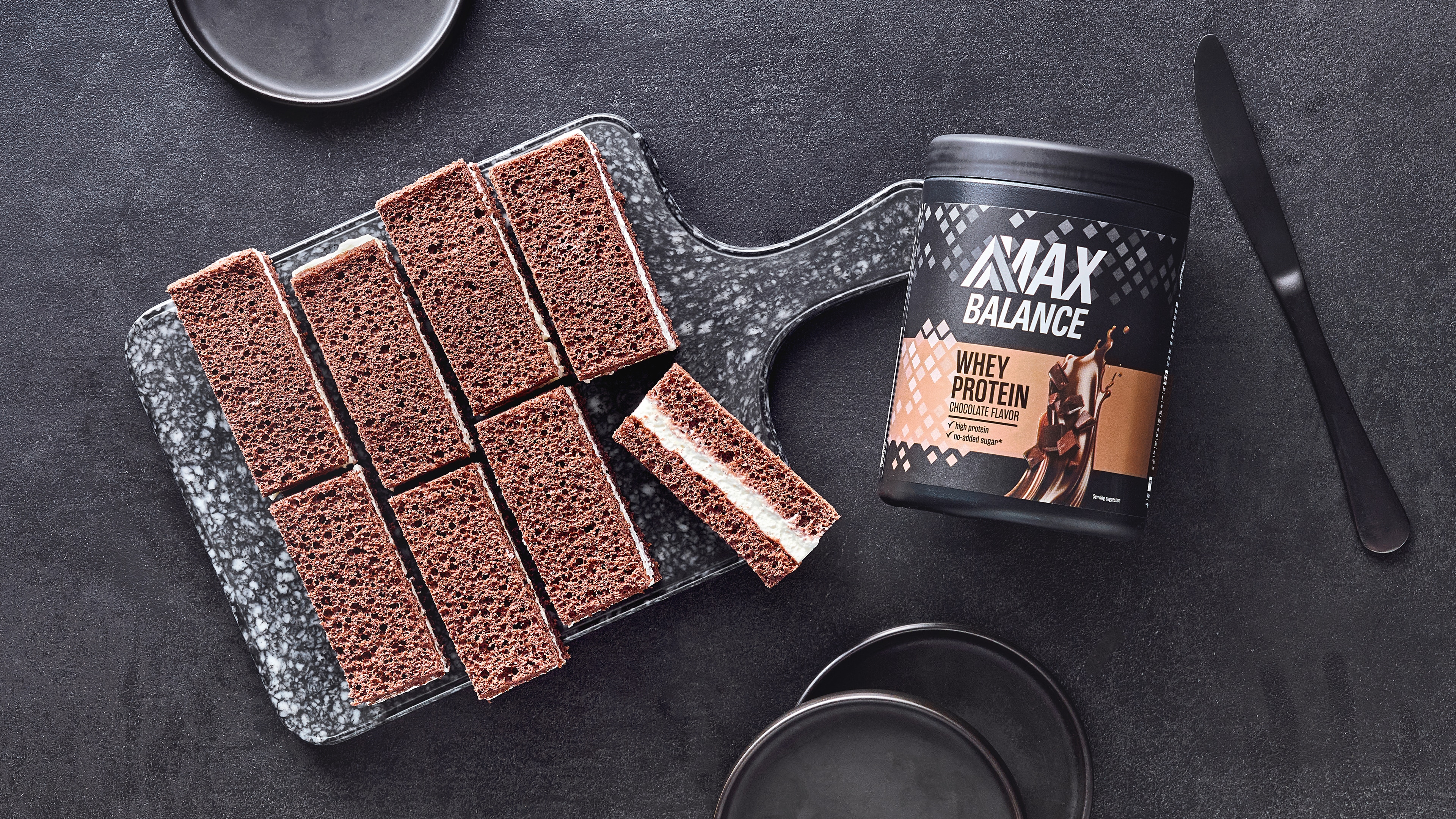 Teigige mit heller Creme gefüllte dunkle Schnitten auf Brettchen, daneben eine Dose MaxBalance Whey Proteinpulver Schokolade