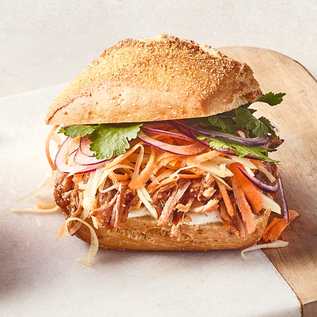 Ein Teller mit einem Sandwich aus hellem Brötchen, gefüllt mit gezupftem Fleisch, roten Zwiebelringen, Karottenstreifen, Weißkohl und Koriander.