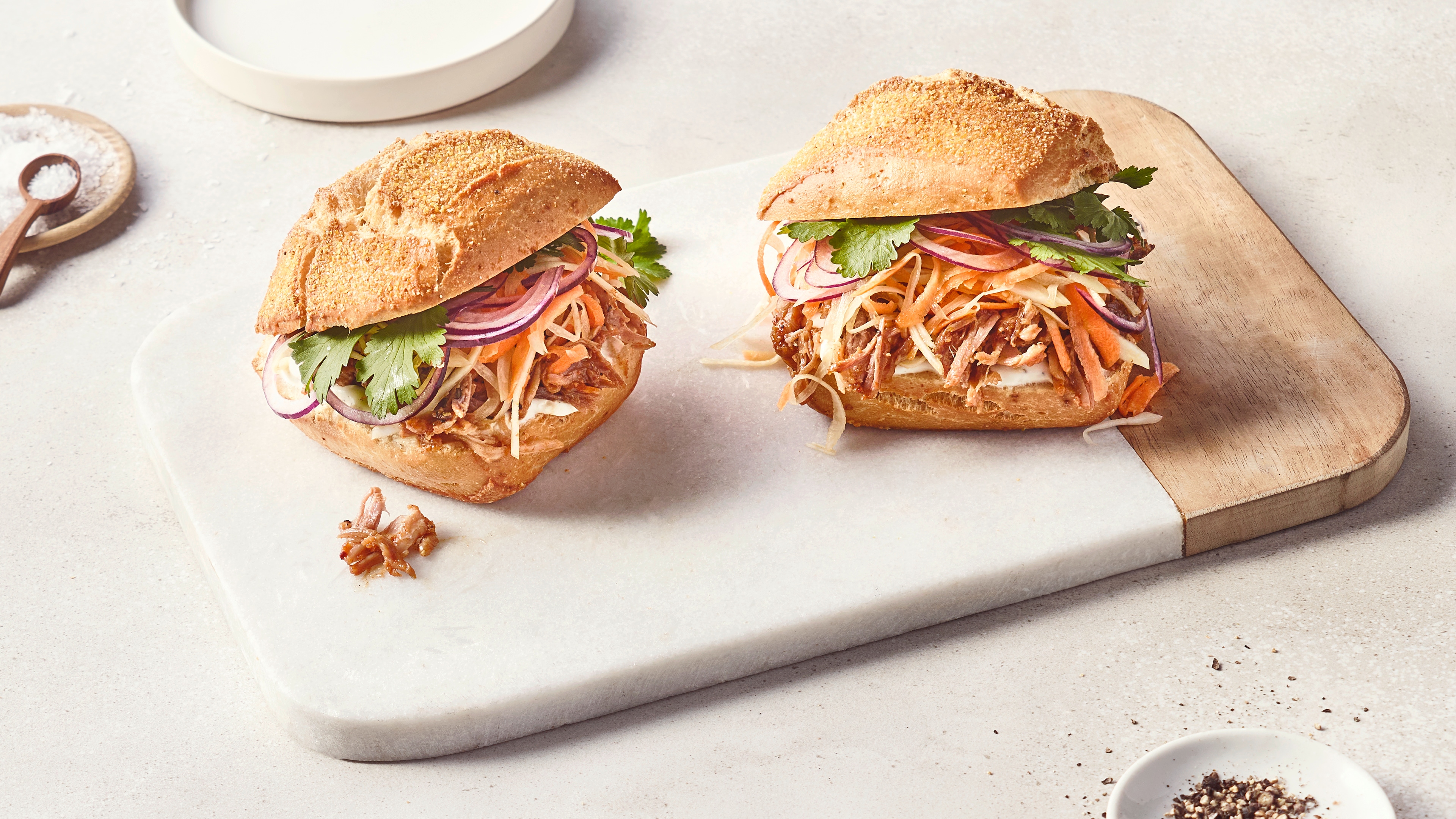 Ein Teller mit einem Sandwich aus hellem Brötchen, gefüllt mit gezupftem Fleisch, roten Zwiebelringen, Karottenstreifen, Weißkohl und Koriander.