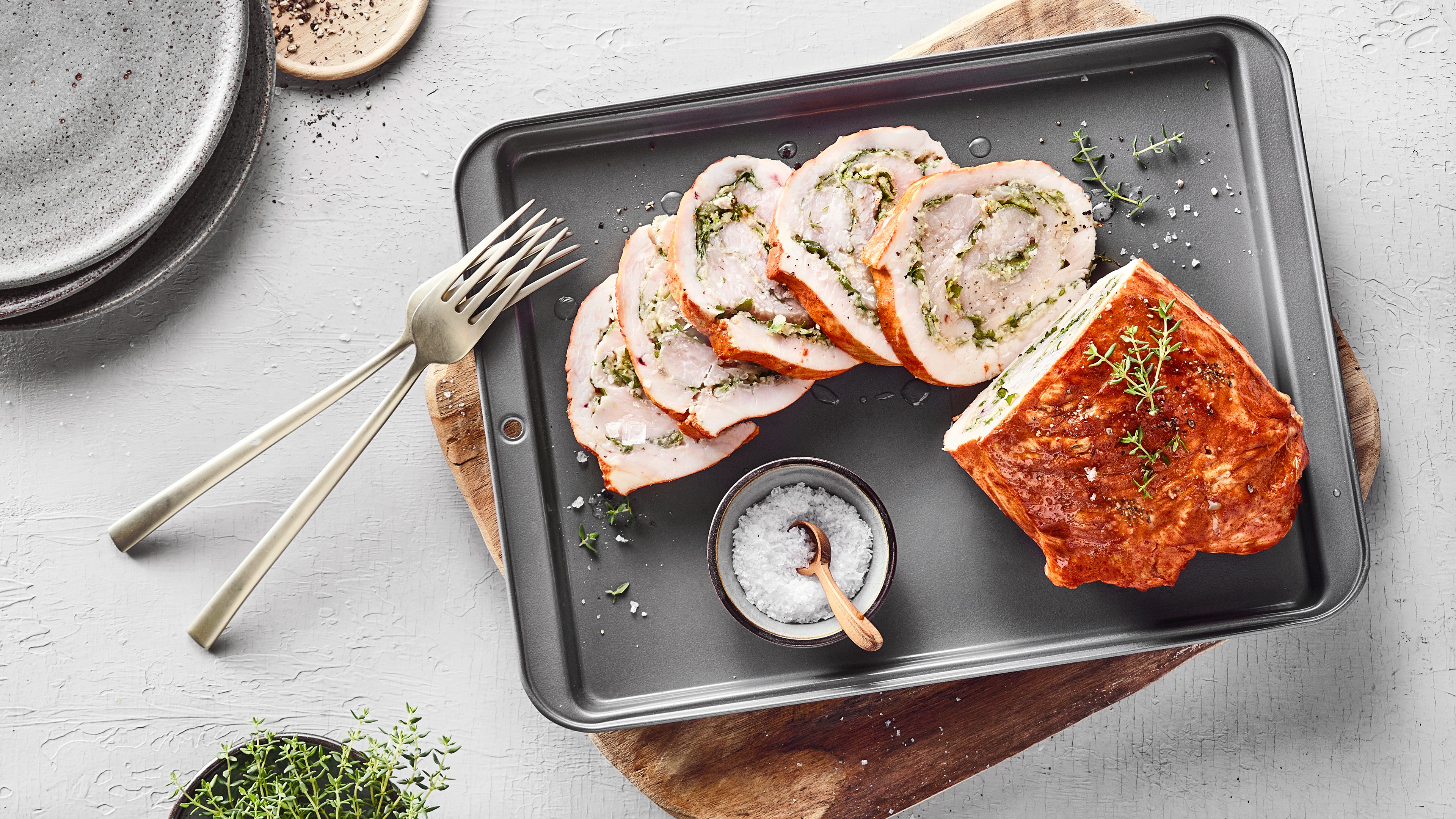 Auf einem Blech liegen Scheiben eines gefüllten Bratens mit Thymian, daneben Salzschale und Gabeln.