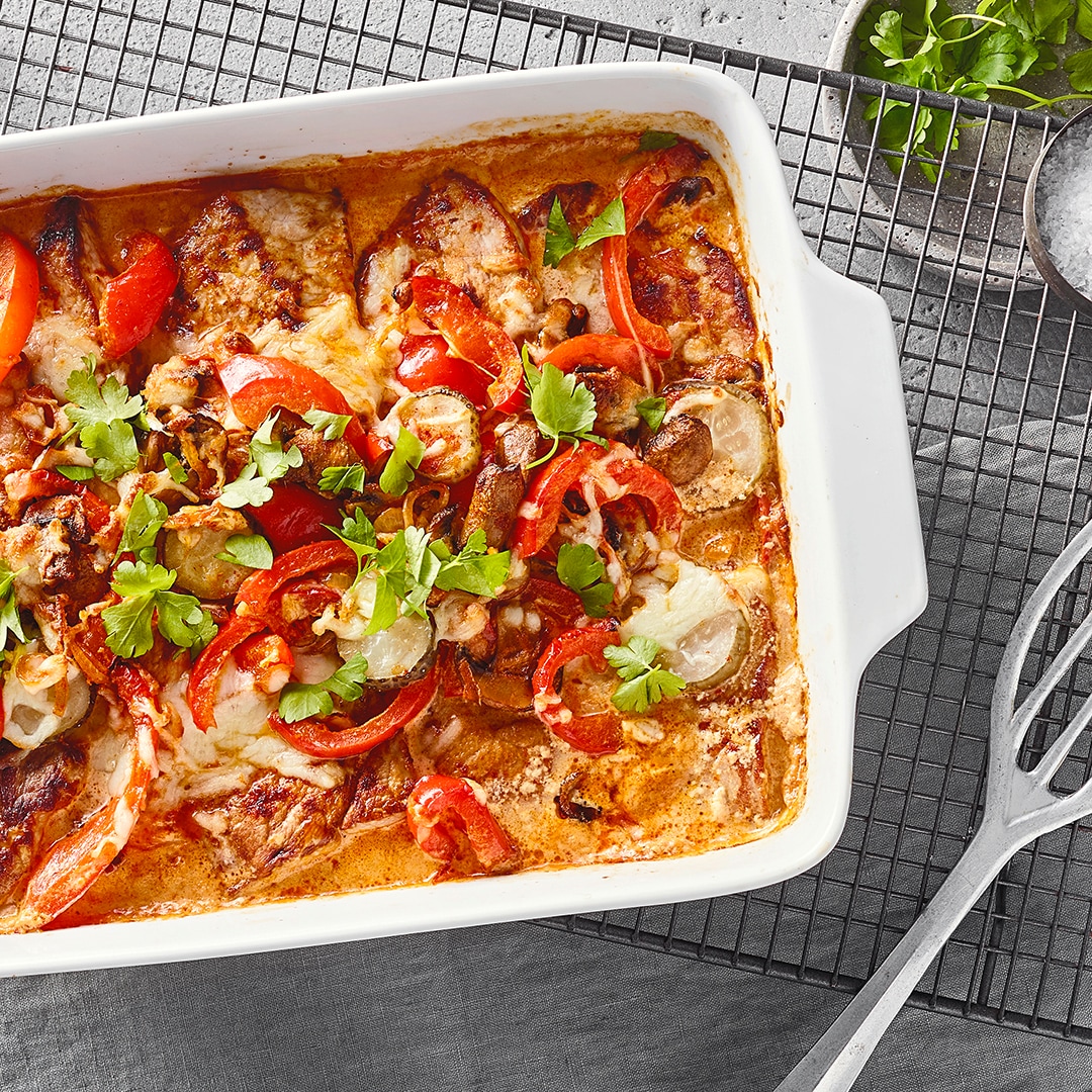 Eine Auflaufform mit Fleisch, roter Paprika & Gurkenscheiben in roter Soße, mit Käse überbacken, garniert mit Kräutern