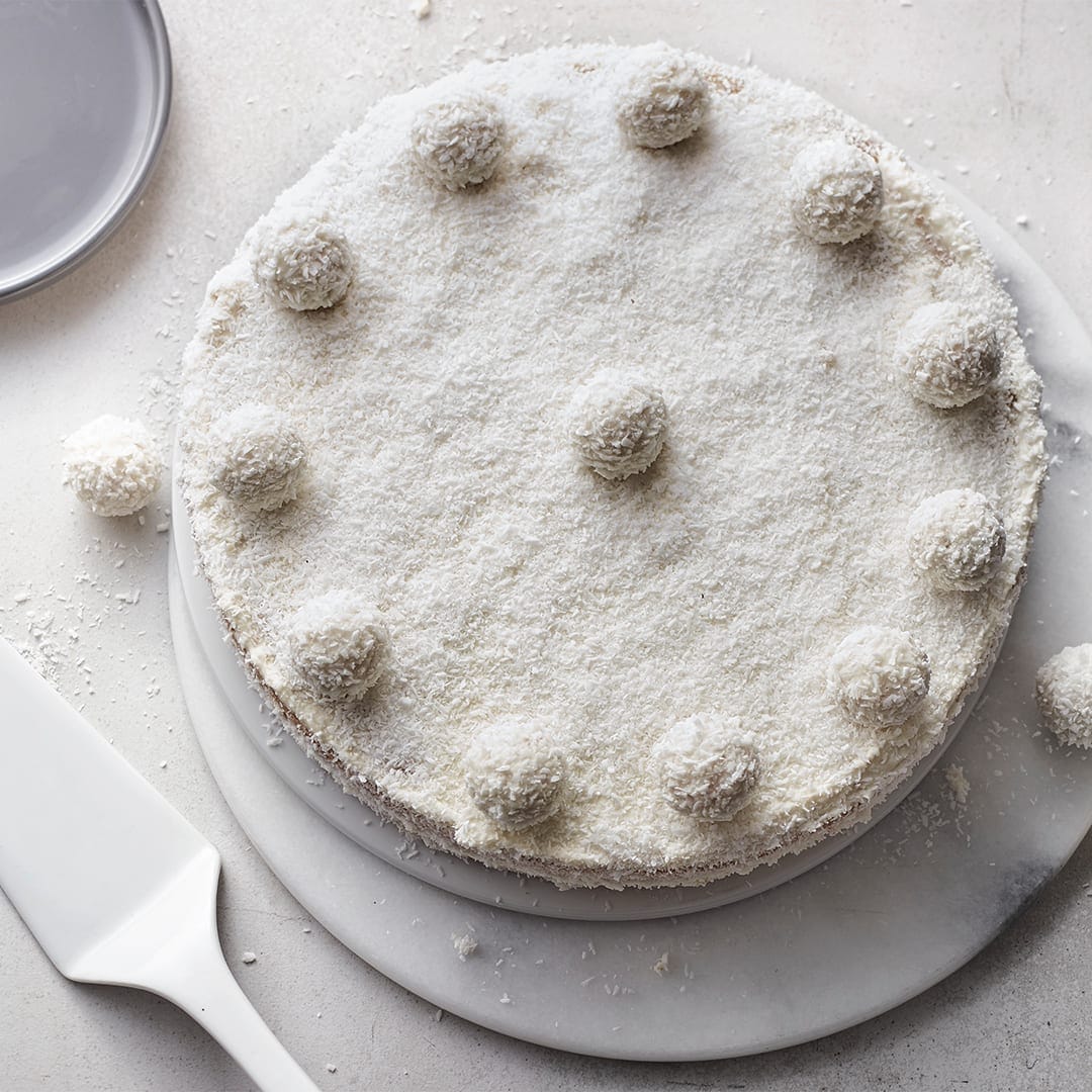 Eine weiße Torte, bedeckt mit Kokosraspeln, getoppt mit Raffaello-Kugeln, auf einer Stein-Marmorplatte