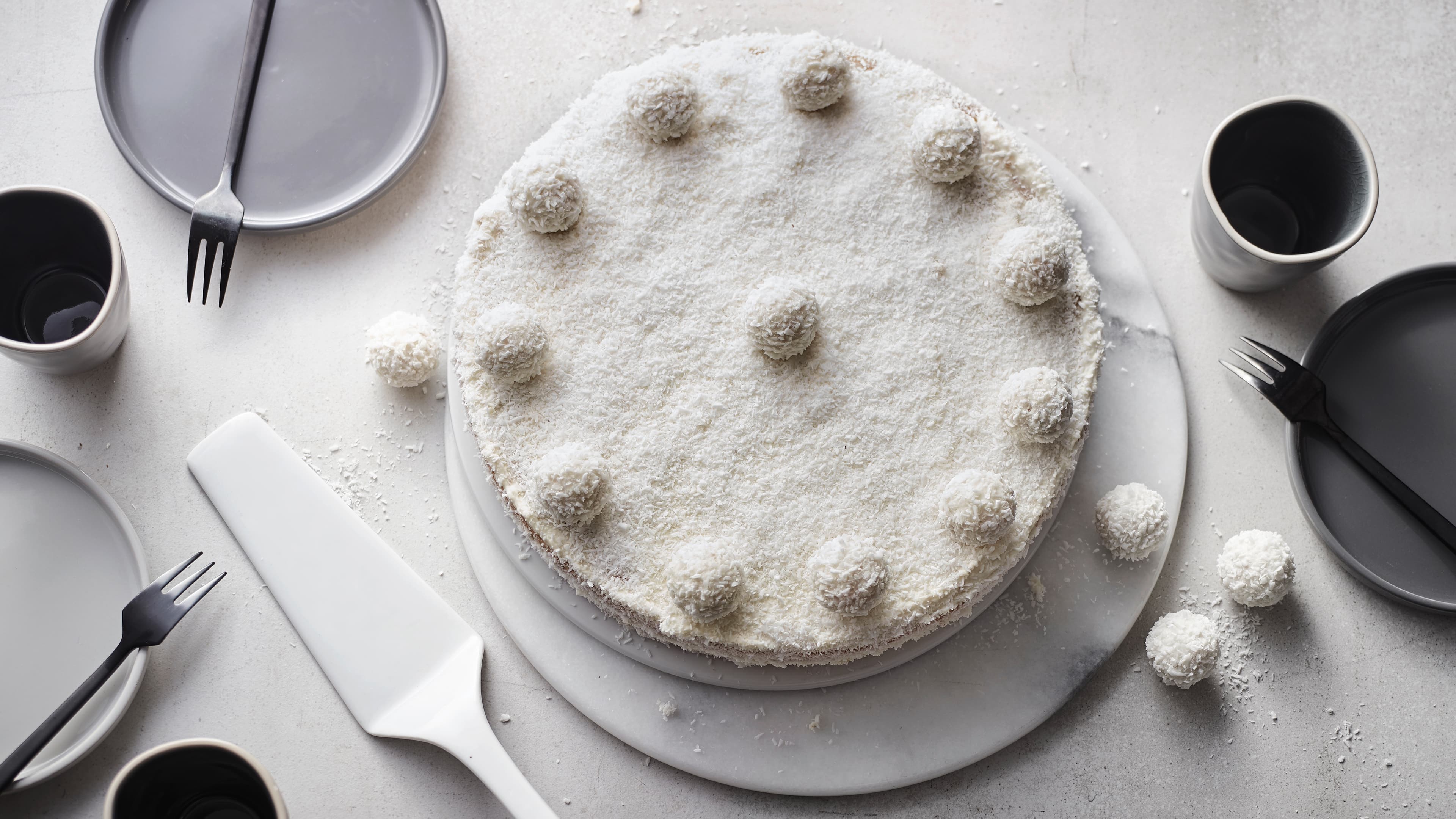 Rezept für Raffaello-Torte Eine weiße Torte, bedeckt mit Kokosraspeln, getoppt mit Raffaello-Kugeln, auf einer Stein-Marmorplatte