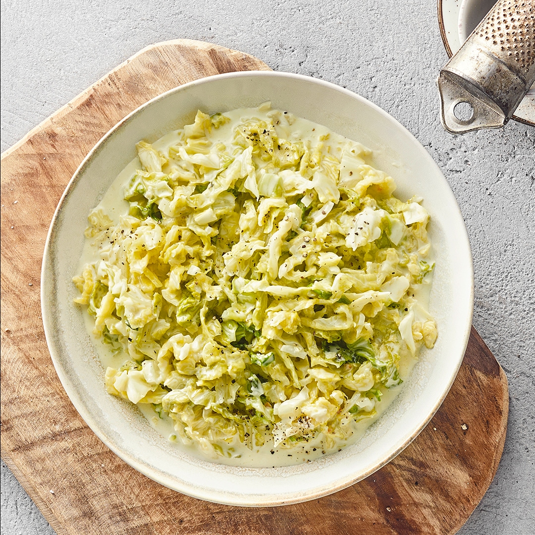 Holzbrett mit einer weißen Schale gefüllt mit Wirsing‑Rahm‑Gericht. Oben rechts liegt eine Muskatreibe.