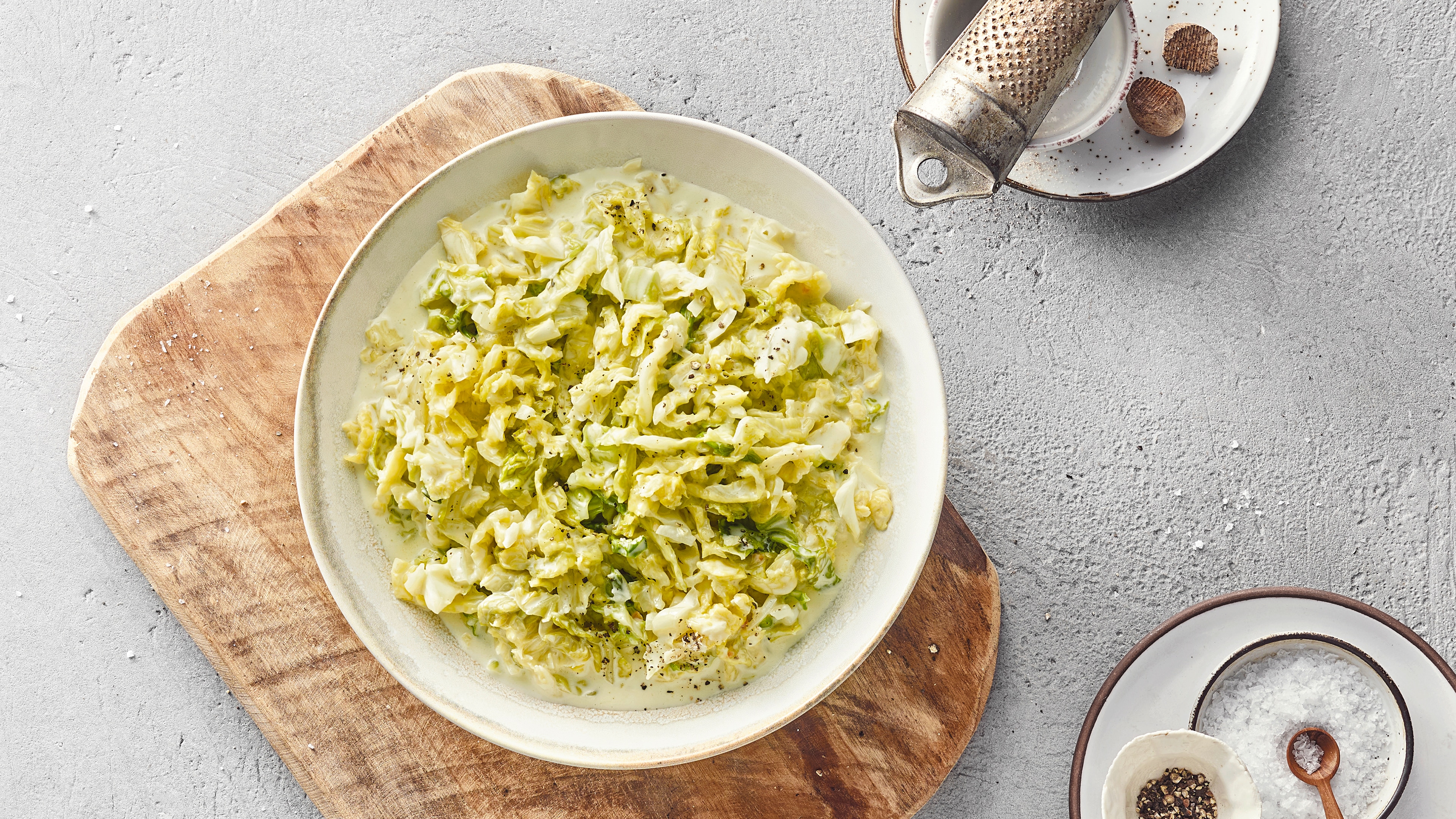 Holzbrett mit einer weißen Schale gefüllt mit Wirsing‑Rahm‑Gericht. Oben rechts liegt eine Muskatreibe.