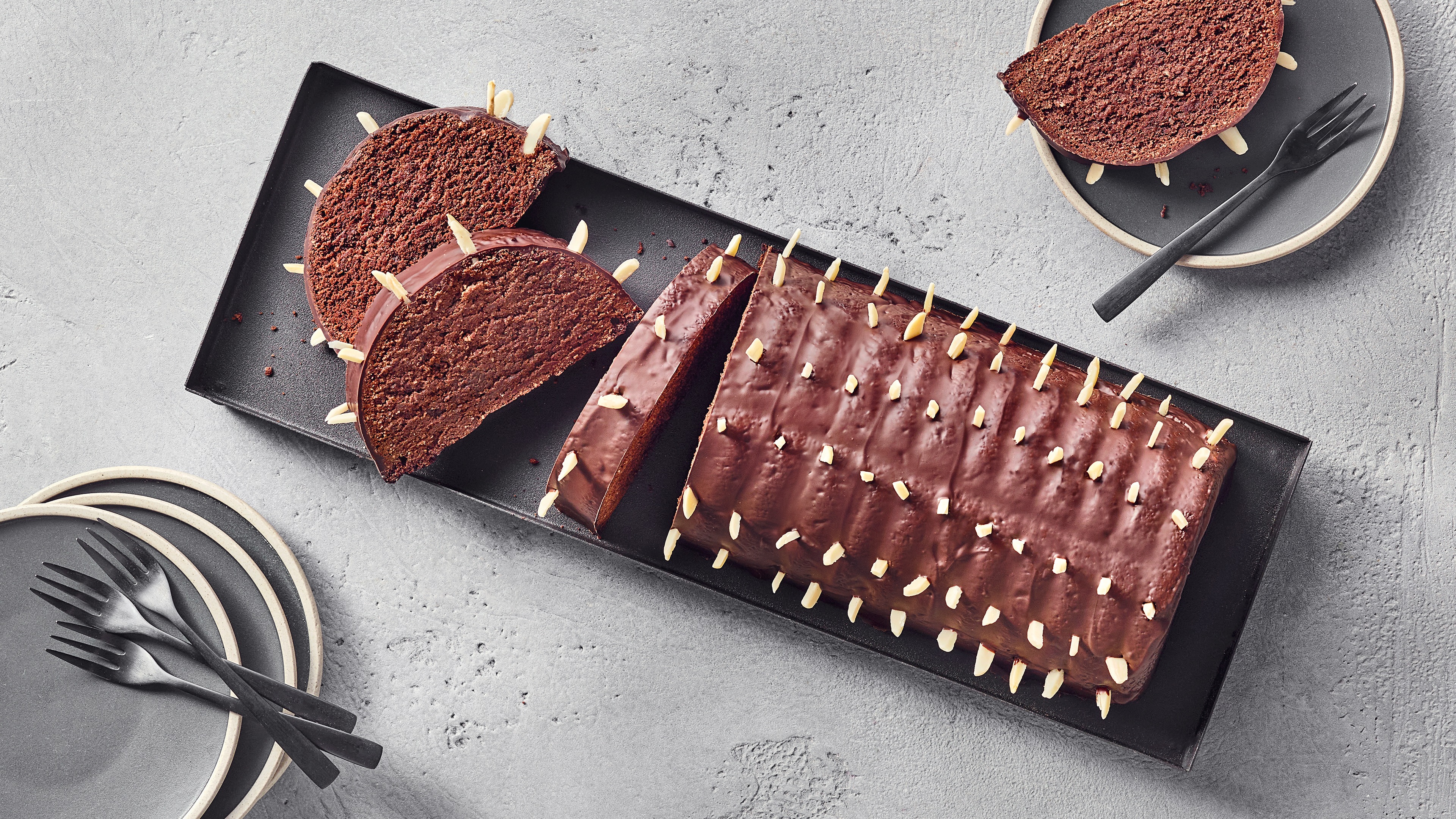 Ein Teller mit dunklem Schokokuchen, überzogen mit glänzendem Guss und bestreut mit hellen Mandelstiften, in Scheiben geschnitten.