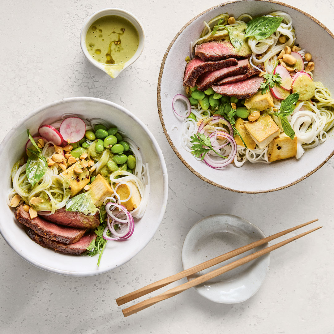 Zwei Schalen mit Reisnudeln, rötlichen Rindfleischscheiben, Tofu, Edamame und Soße.