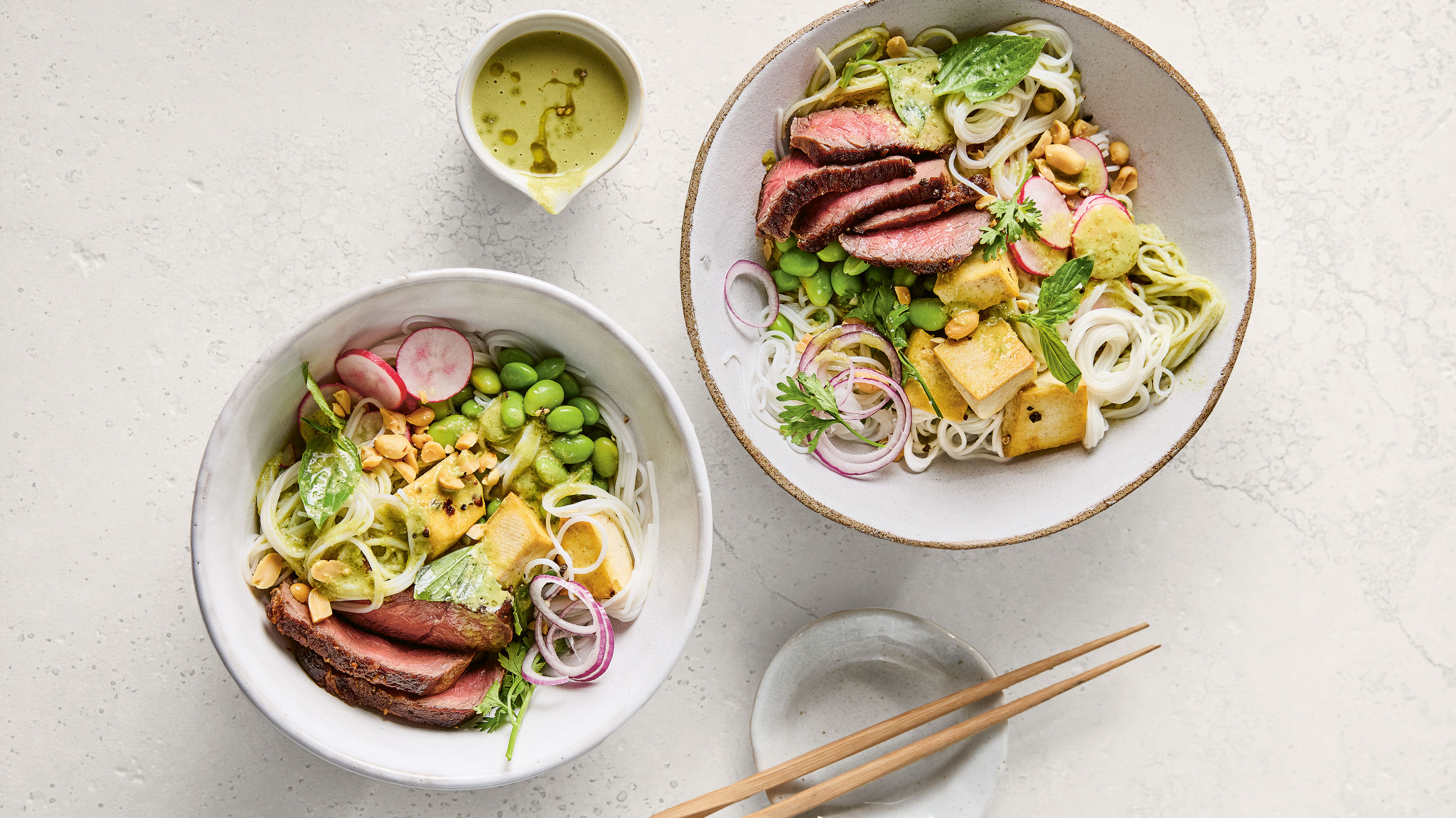 Zwei Schalen mit Reisnudeln, rötlichen Rindfleischscheiben, Tofu, Edamame und Soße.