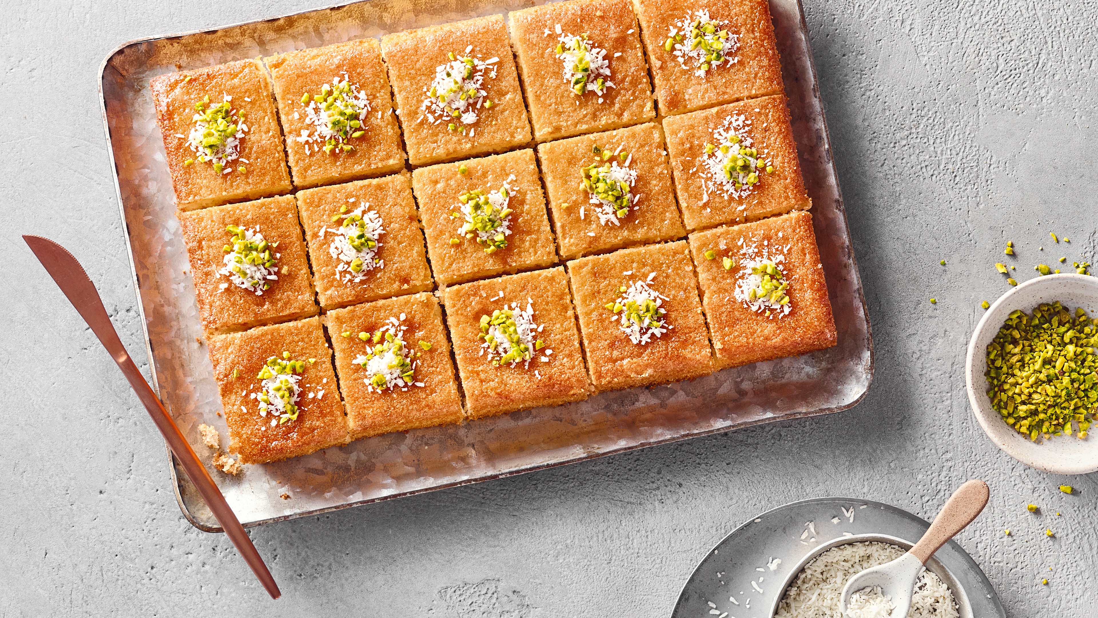 Quadratische Grießkuchenstücke auf Blech. Daneben ein Schälchen Pistazien.