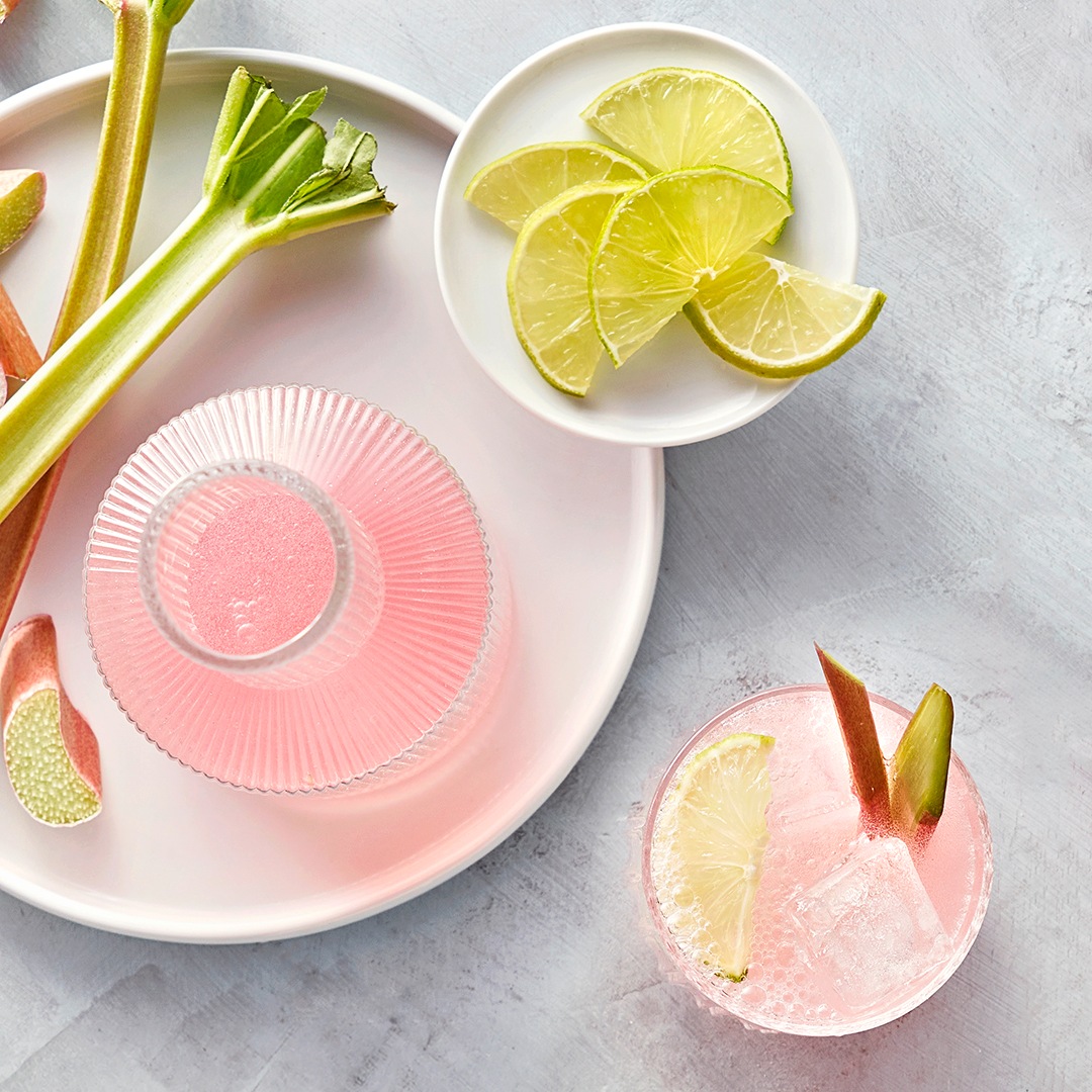 Hellrosa Flüssigkeit in Flasche und Glas, daneben Rhabarberstangen und Limettenscheiben