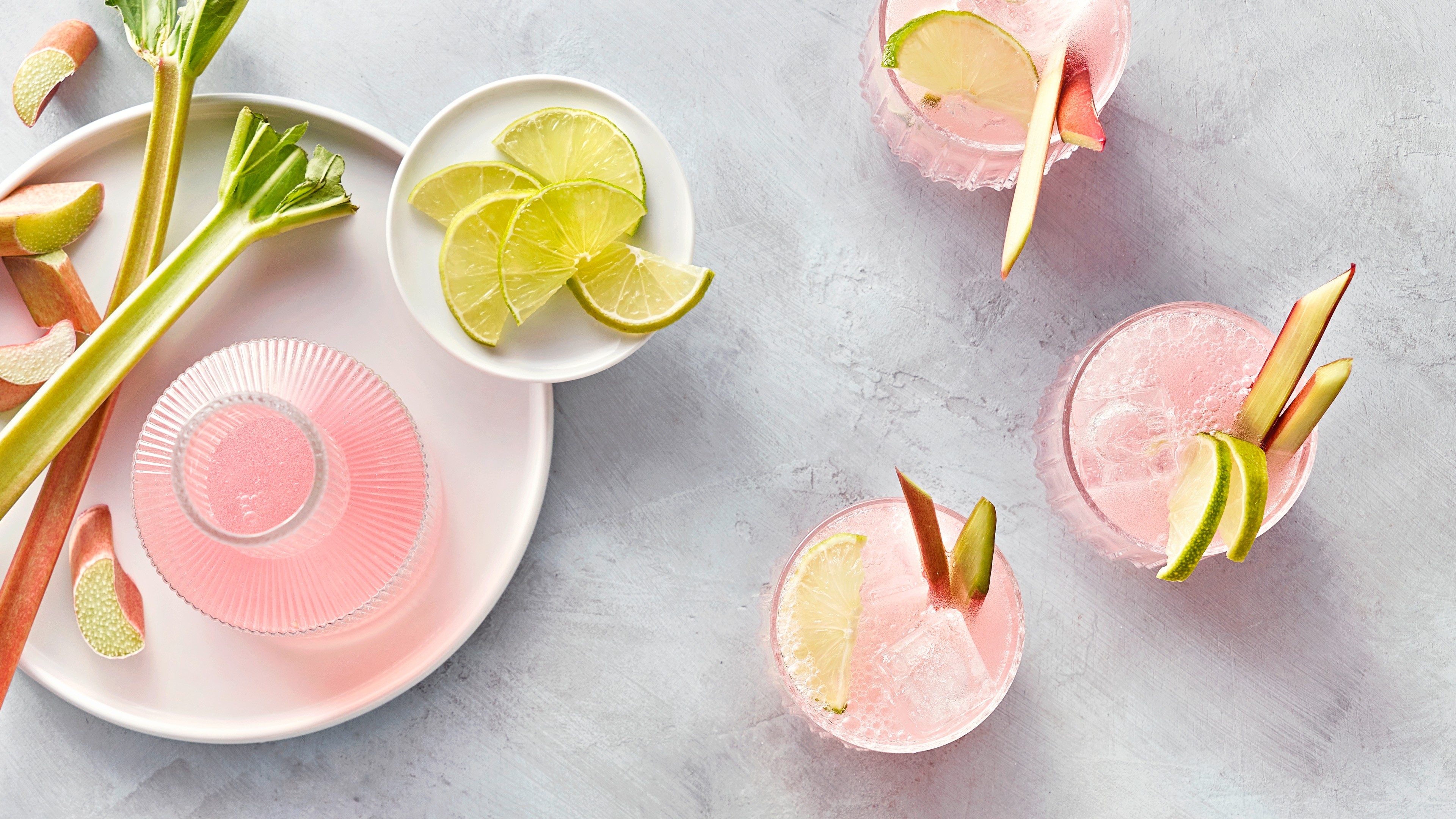 Hellrosa Flüssigkeit in Flasche und Gläsern, daneben Rhabarberstangen und Limettenscheiben