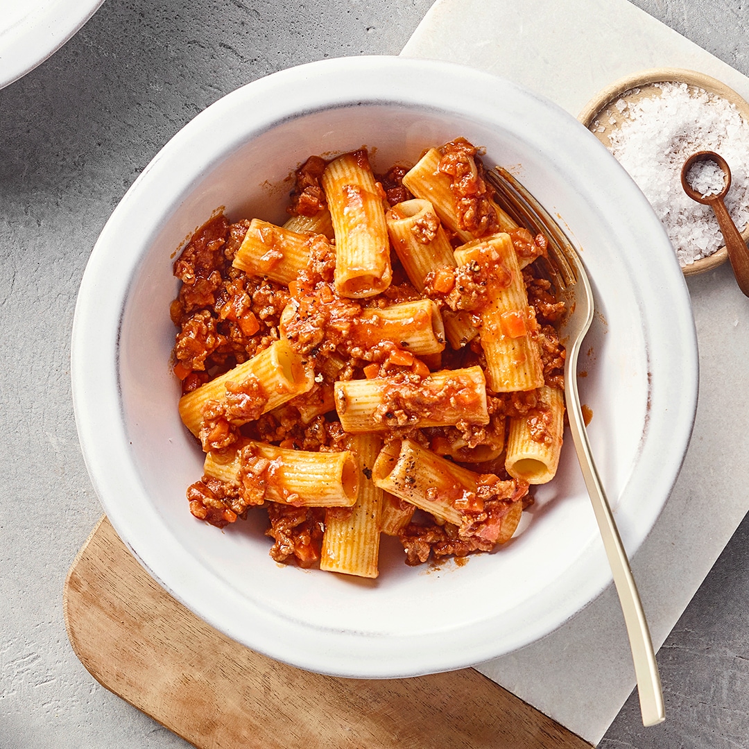 Ein Teller mit Rigatoni in roter Hackfleischsoße, darin kleine orange Gemüsestücke, daneben eine Gabel.