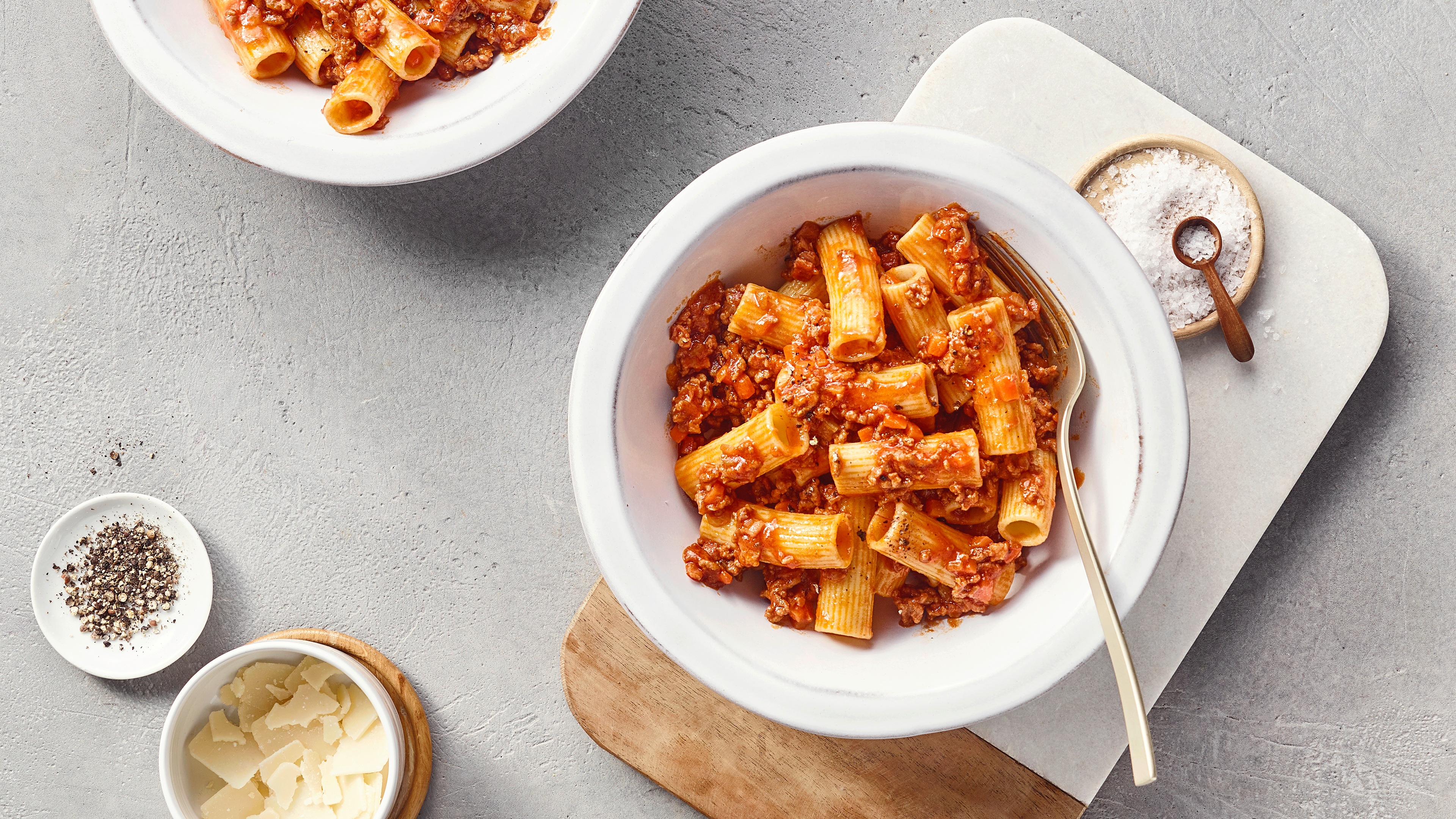 Ein Teller mit Rigatoni in roter Hackfleischsoße, darin kleine orange Gemüsestücke, daneben eine Gabel.