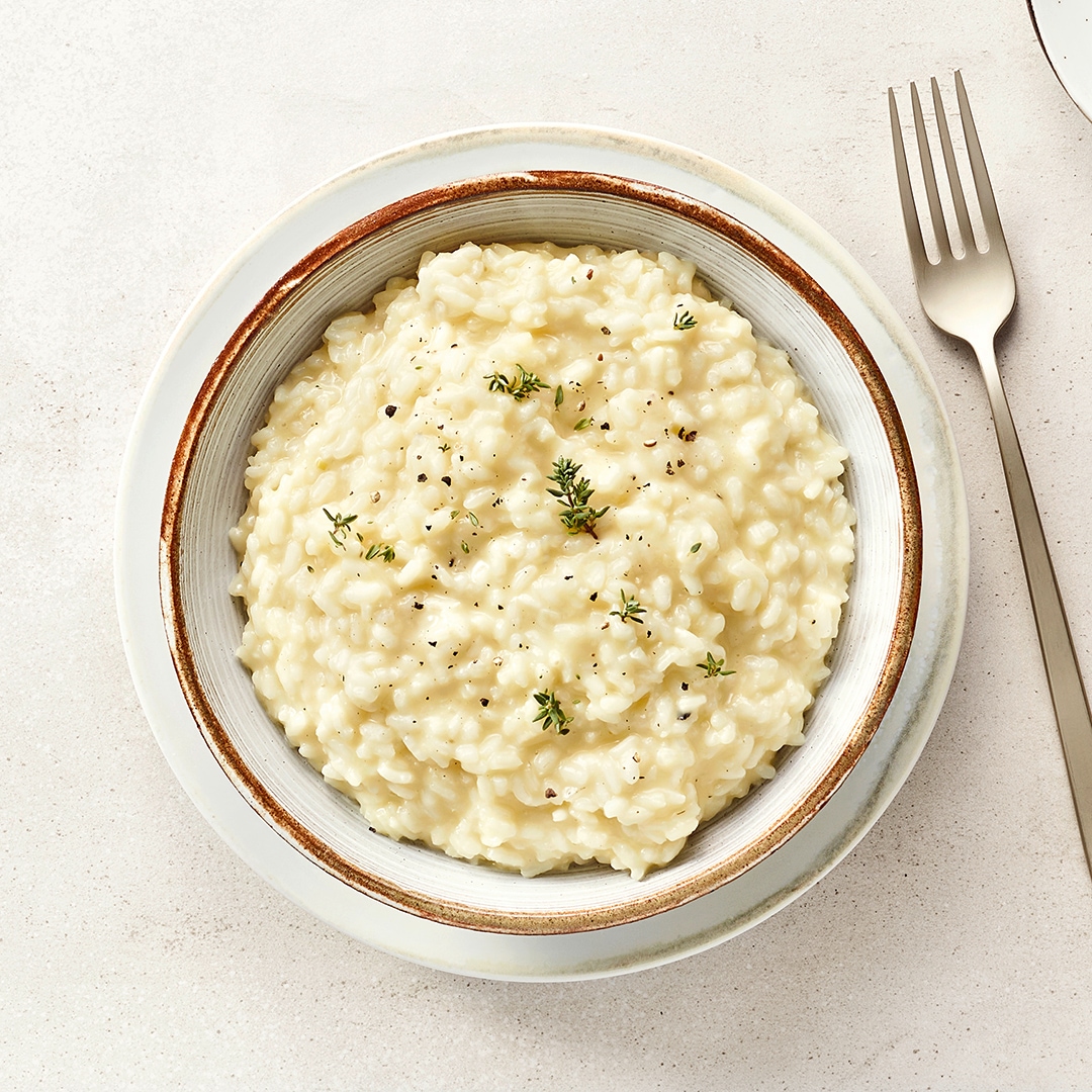 Ein Teller mit weißem cremigem Risotto, bestreut mit schwarzen Pfefferkörnern und frischen Kräutern.
