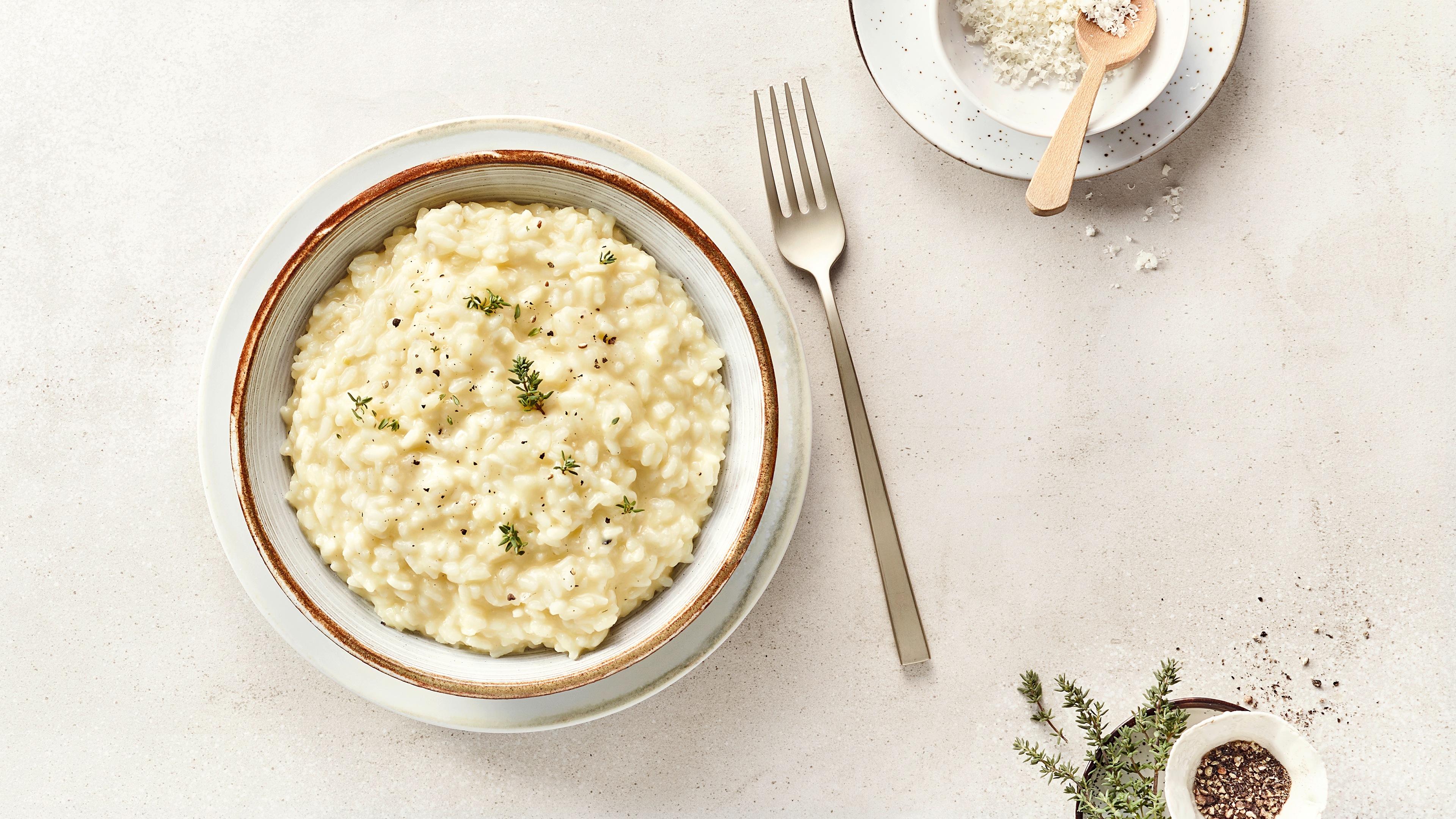 Ein Teller mit weißem cremigem Risotto, bestreut mit schwarzen Pfefferkörnern und frischen Kräutern.