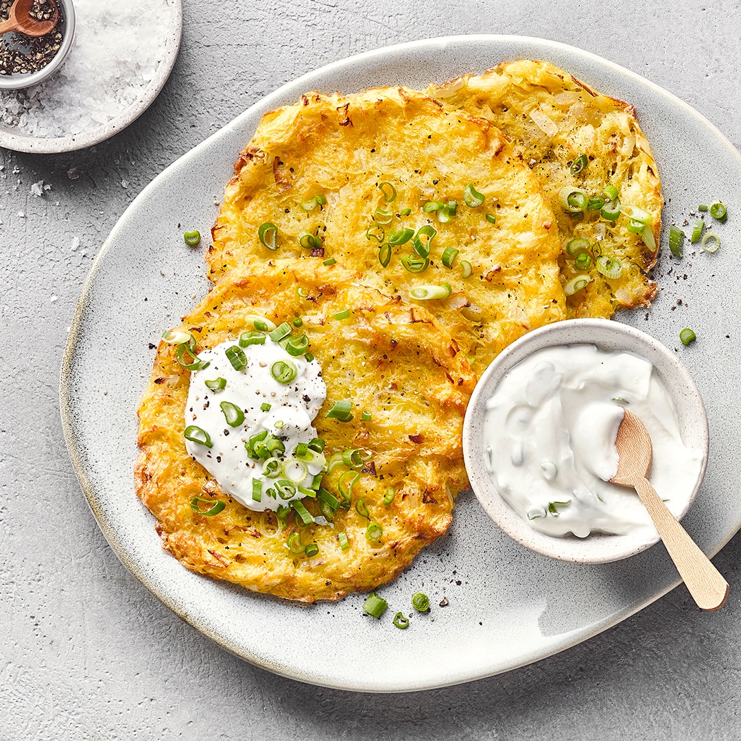 Unregelmäßig runder Teller mit drei Röstis, Sourcream und Frühlingszwiebeln. Auf dem Teller steht zusätzlich eine kleine Schale Sourcream mit einem Holzlöffel. Oben links befindet sich eine Schale mit Salz.