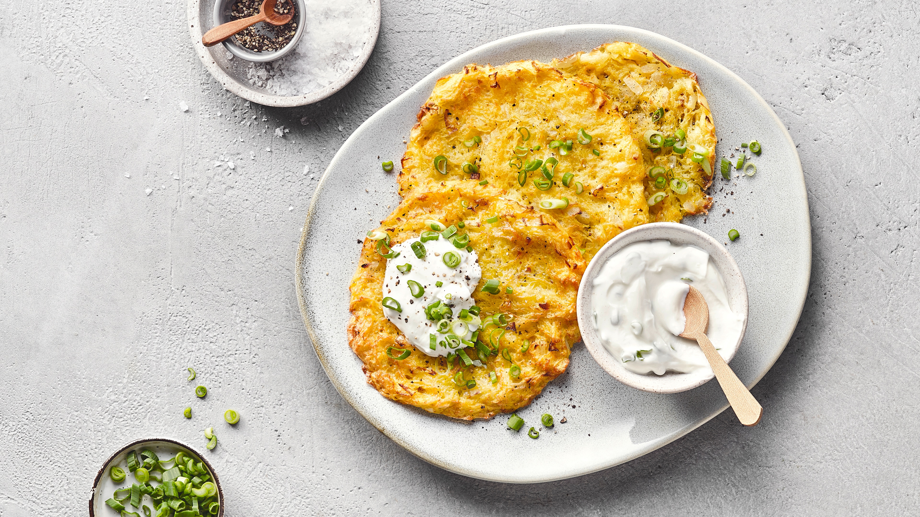 Unregelmäßig runder Teller mit drei Röstis, Sourcream und Frühlingszwiebeln. Auf dem Teller steht zusätzlich eine kleine Schale Sourcream mit einem Holzlöffel. Oben links befindet sich eine Schale mit Salz, unten links eine Schale mit Frühlingszwiebeln.