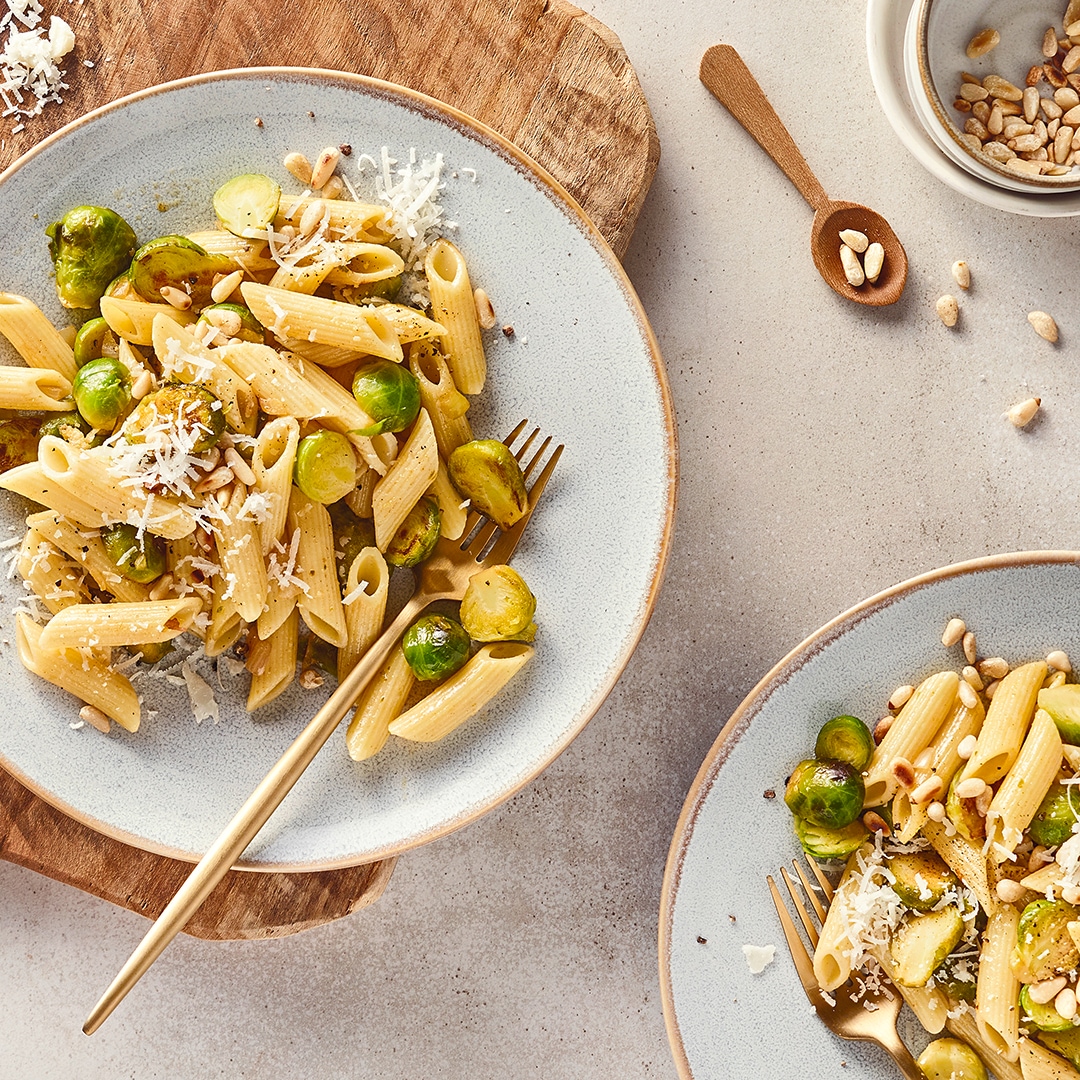 Runde Teller mit Penne-Pasta, getoppt mit geröstetem Rosenkohl, geriebenem Hartkäse und gerösteten Pinienkernen
