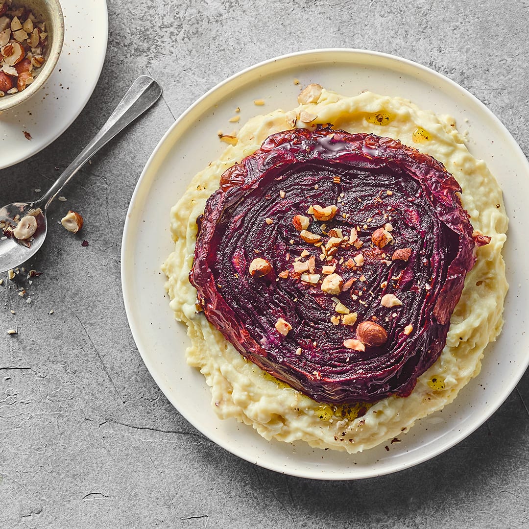 Ein saftiges Rotkohl-Steak, gebettet auf cremigem Kartoffelstampf, getoppt mit Haselnüssen, auf einem runden Teller