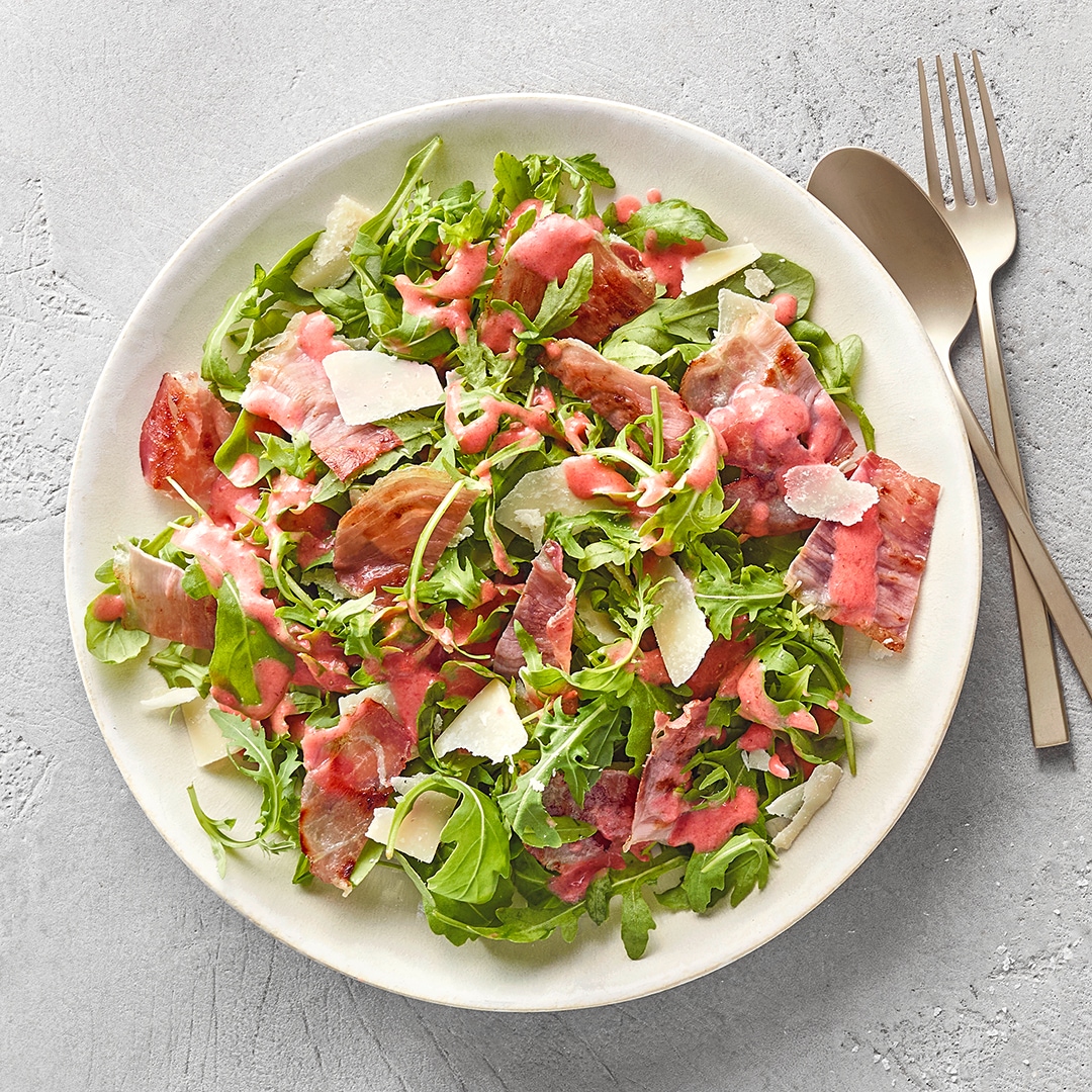 Blattsalat mit knusprigem Speck, Parmesanspänen und rosa Dressing auf einem weißen Teller, daneben Besteck.
