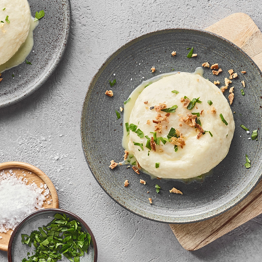 Ein Teller mit einer runden weißen Dampfnudel, darauf grüne Kräuter und braune Krümel.